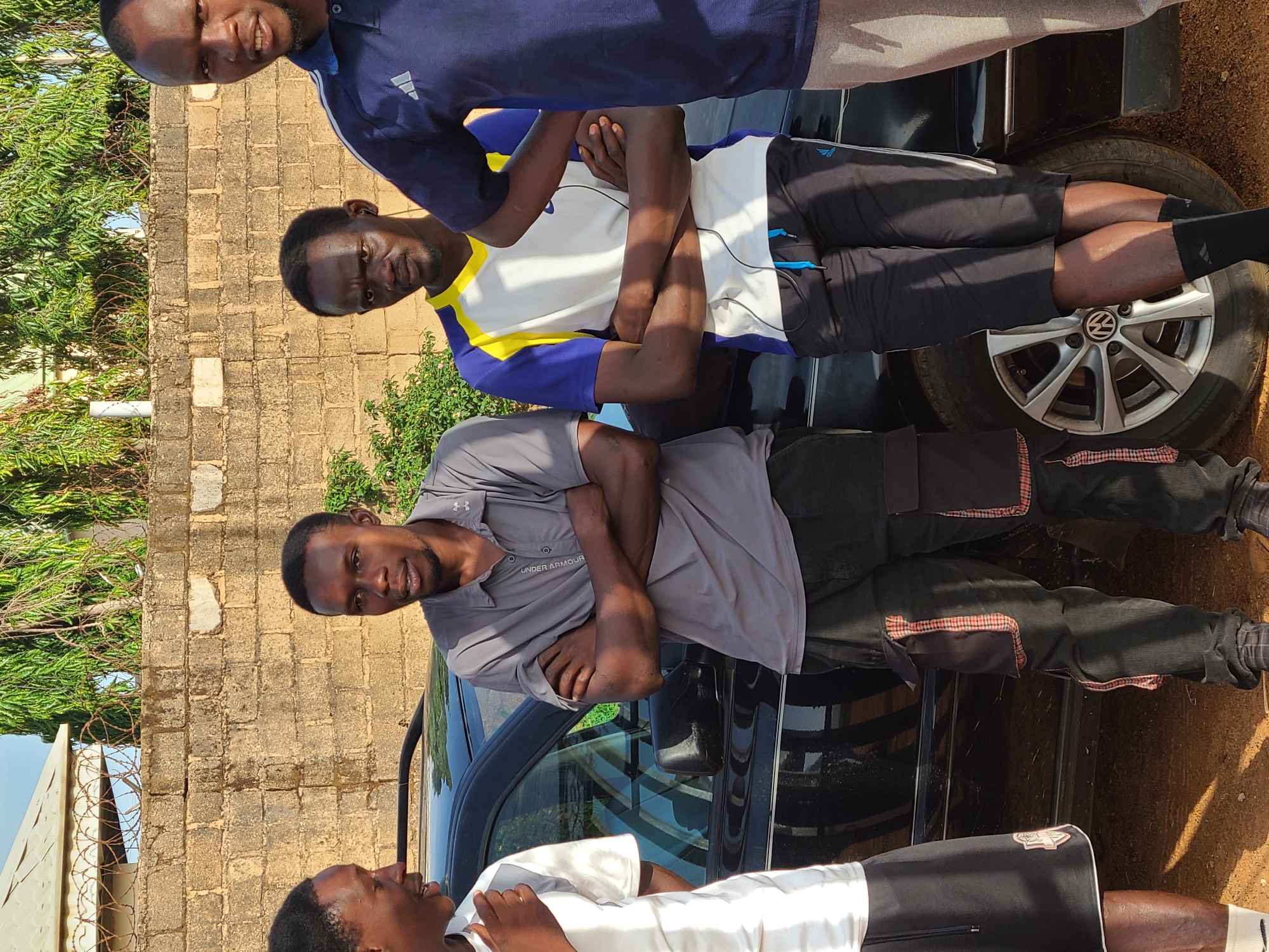 Four adult males are outside in Jos, Nigeria. Three of the men stand behind and near a black car. The car’s rear driver side tire is visible with a Volkswagen logo on the wheel. One man wears a gray collared shirt, another wears a white shirt with blue and yellow accents, and a third wears a blue collared shirt with an Adidas logo. The fourth man is partially visible in the foreground and wears a white shirt. They all have their arms crossed. A brick wall, trees, and a building are in the background.