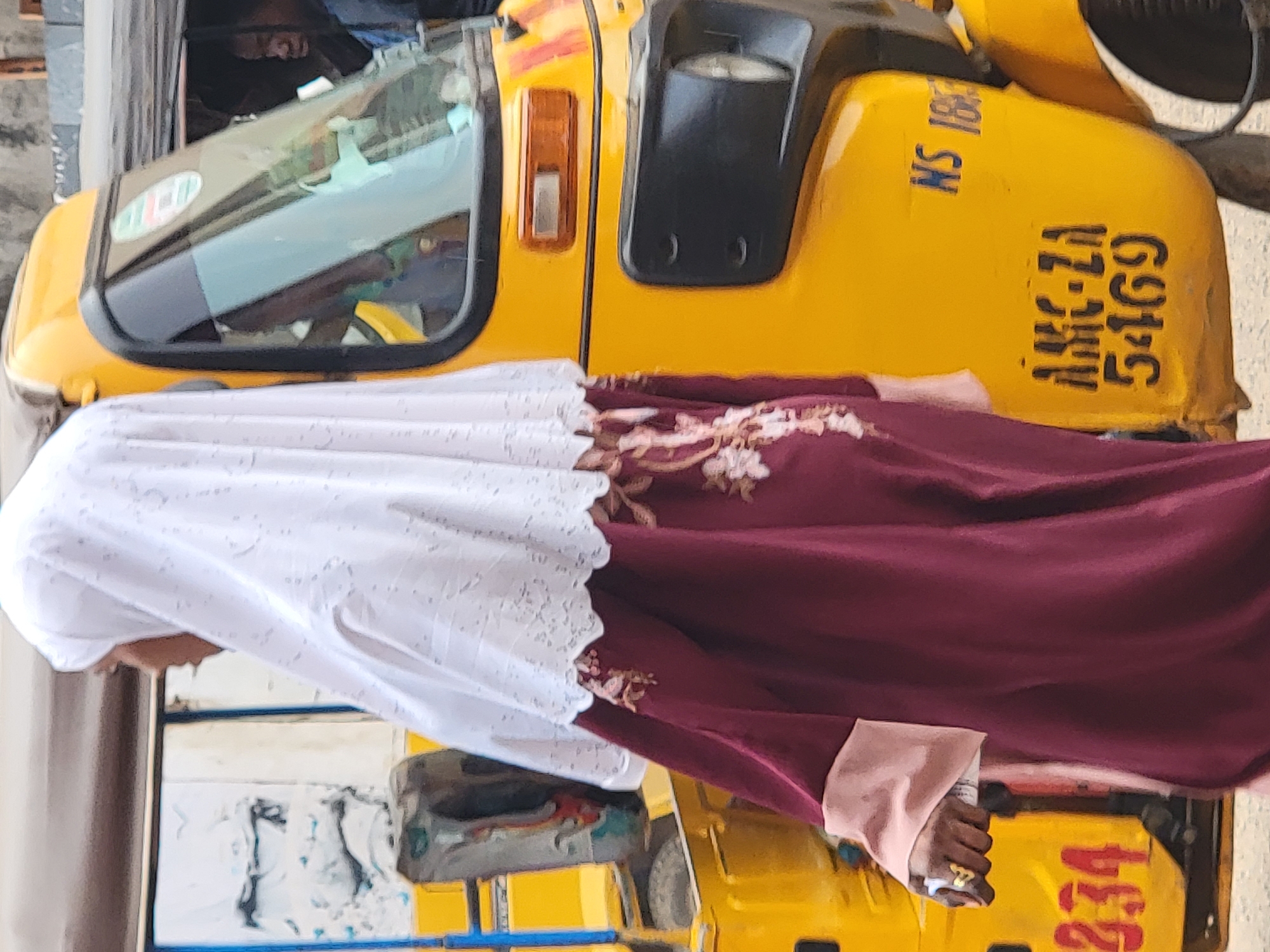 A person wearing a white and burgundy dress stands in front of a yellow "Keke Napep" tricycle taxi in Maiburiji, Nigeria. The taxi has the license plate "AMC-ZA 5169" and the number "234". A person can be seen inside the taxi.