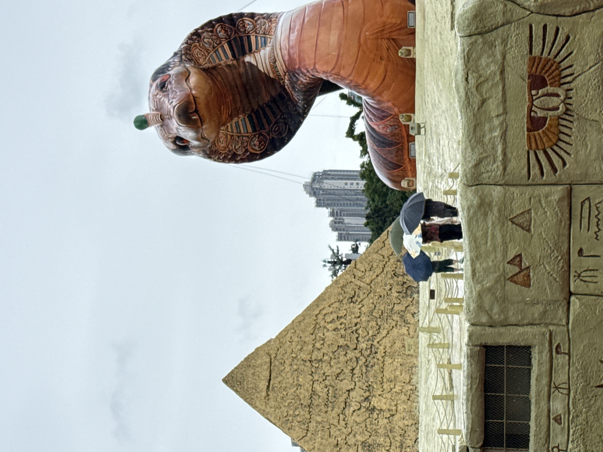 In the city of Nanshan District, China, on a cloudy day, a large, inflatable cobra sculpture dominates the upper right of the frame, its head turned towards the viewer and a small green ornament on its forehead. To the left of the cobra, a golden pyramid structure is visible. In the foreground, a beige, textured wall is decorated with Egyptian hieroglyphs, including a winged sun disc motif. Three individuals stand on a raised platform behind this wall, holding umbrellas against the rain. They are positioned between the pyramid and the cobra sculpture. In the background, buildings rise above a line of trees. The ground appears wet, suggesting recent rainfall.