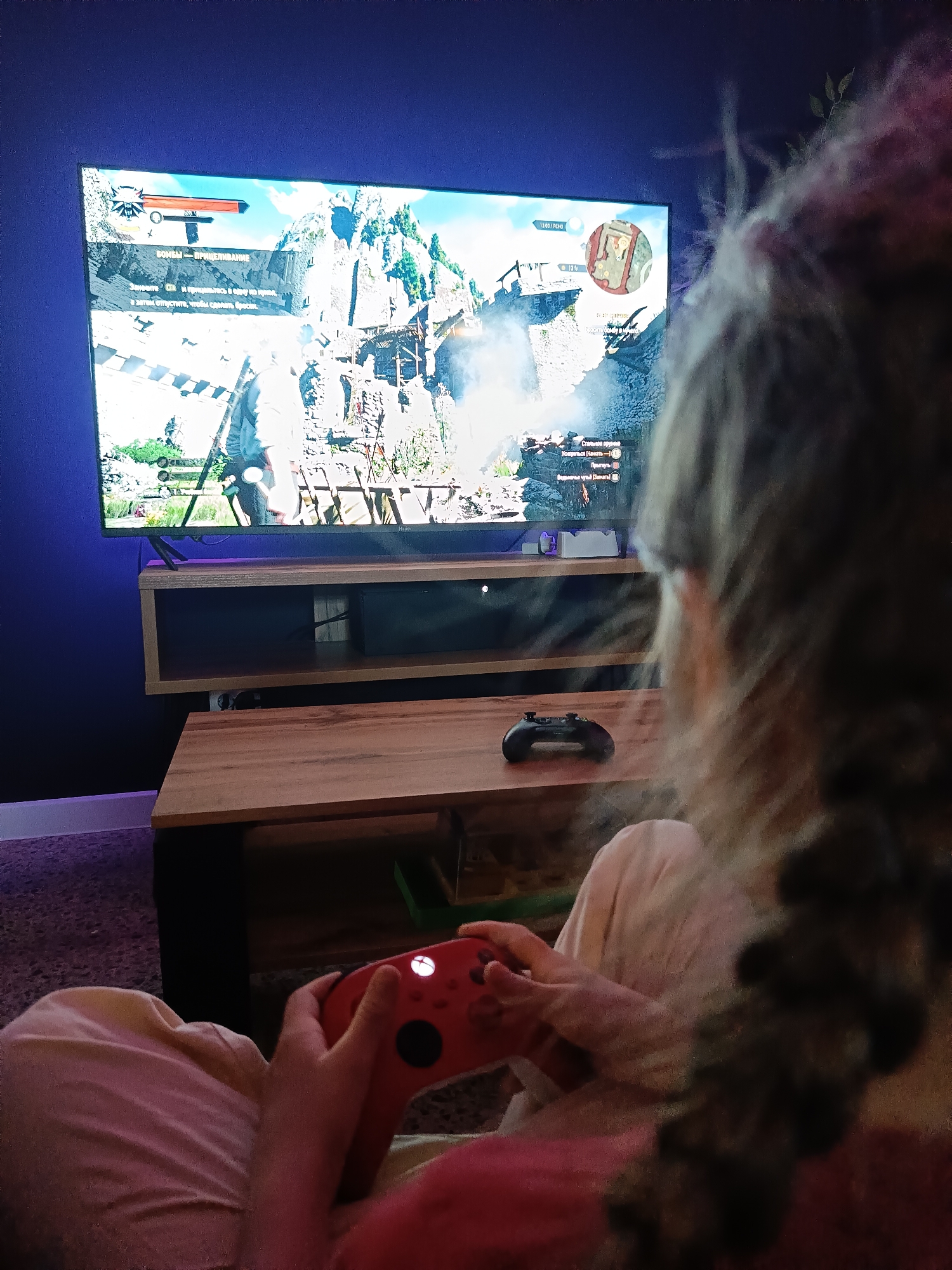 An indoor scene, likely in Chaikovsky, Russia, shows a person playing a video game in dim lighting, suggesting evening or night. The person, seen from behind with braided hair, is holding a red Xbox controller and appears engrossed in the game. A second black controller rests on a wooden table in front of them.
A large flat-screen TV displays gameplay from what appears to be "The Witcher 3: Wild Hunt." The game shows a rugged, mountainous landscape with medieval-style structures and a character in the foreground, likely Geralt of Rivia. The game's user interface is visible, including a health bar, a minimap at the top right, and various text prompts in Russian.
Visible text on the screen includes a directive for bomb usage: "БОМБА — ПРИЦЕЛИВАНИЕ" (BOMB — AIMING) and instructions: "Зажмите Q и прицельтесь в одну из бомб, а затем отпустите, чтобы бросить." (Press Q and aim at one of the bombs, then release to throw.) Below, in-game actions are listed: "Стальное оружие" (Steel weapon), "Ускориться (Нажать А)" (Accelerate (Press A)), "Прыгнуть (B)" (Jump (B)), and "Ведьмачье чутье (Нажать)" (Witcher Senses (Press)).
The TV sits on a modern wooden media console. On a shelf below the TV, a black video game console (resembling an Xbox Series X) is visible, along with a white controller charging station. The background features a dark blue wall and a glimpse of a purple/gray carpet.