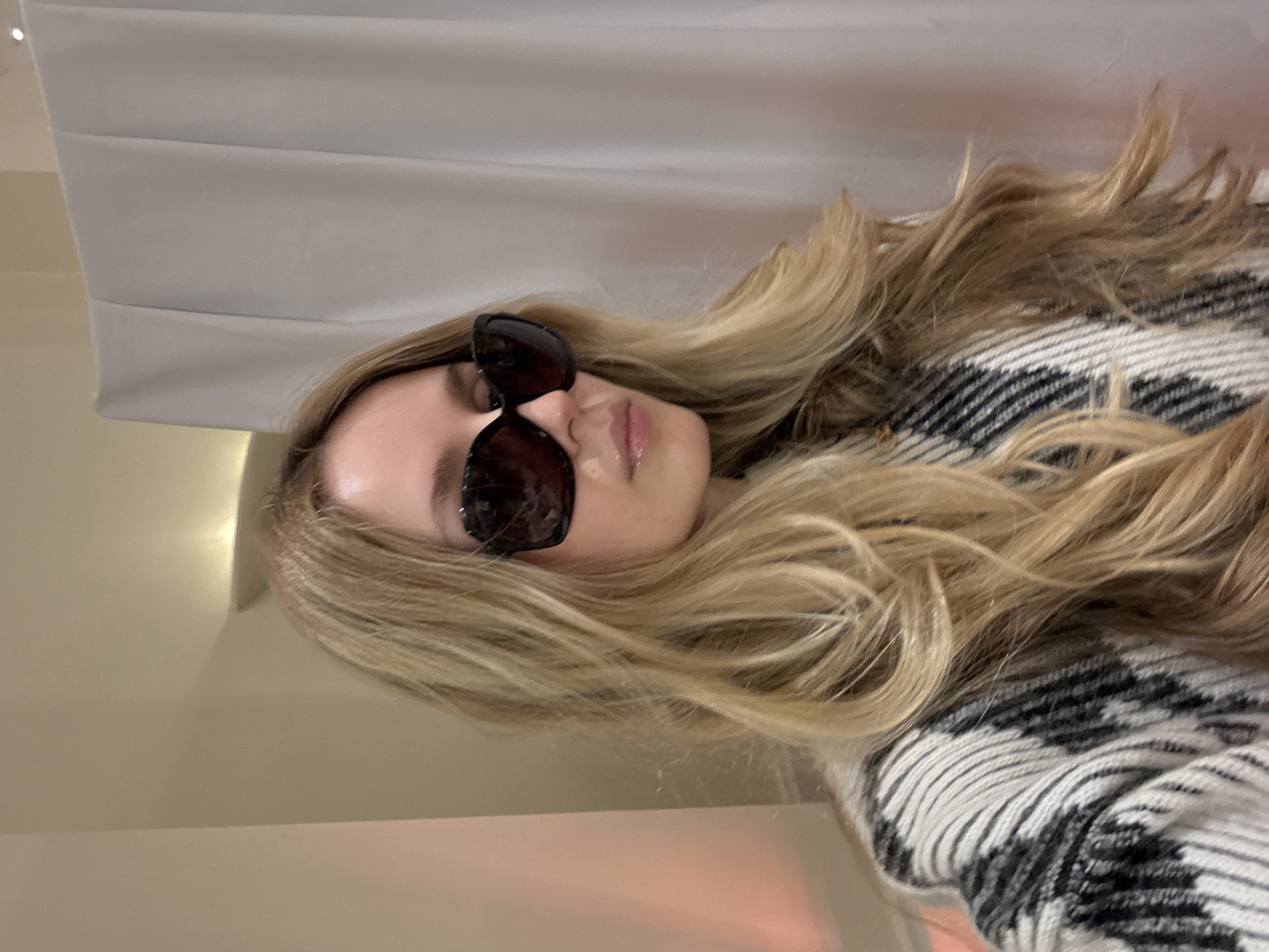 A light-skinned individual with long, blonde wavy hair and sunglasses is partially visible in the image. The person is wearing a white and black patterned garment. A white curtain is visible in the background. The location cannot be determined based on the provided image.