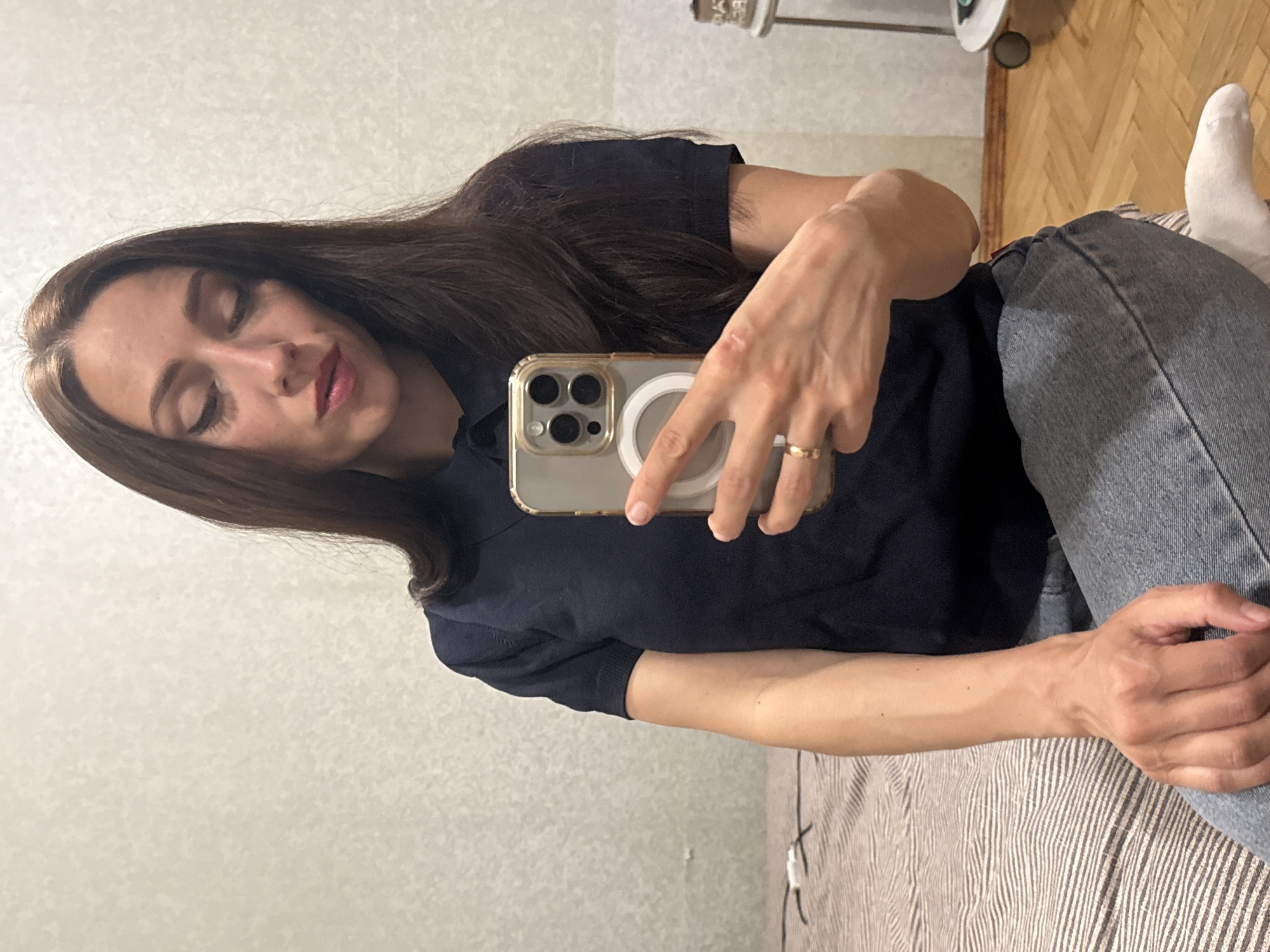 A young adult female is seated on a bed, taking a selfie with her phone. She has long, dark brown hair, fair skin, and is wearing a dark navy polo shirt and light blue jeans. Her eyes are closed, and her head is tilted down, with her lips slightly parted. She is wearing a gold ring on her left ring finger. The bed has a striped comforter. The background appears to be a room with a textured, off-white wall and a wooden floor. A small rolling side table is visible in the background to the right. The overall scene suggests a casual, indoor setting. There is no information in the image to indicate a specific location within Saint Petersburg, Russia.
