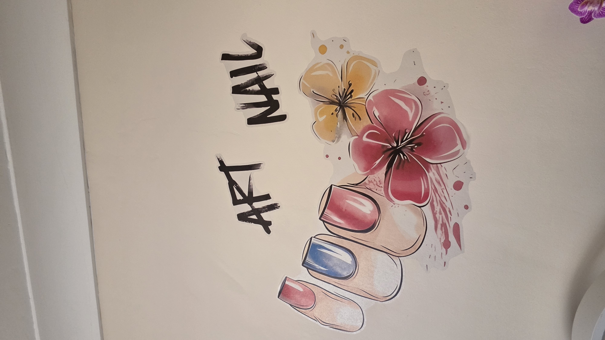 A medium-level, indoor shot presents a light tan wall adorned with a decal featuring the words "ART NAIL" in a stylized, handwritten font, alongside an illustration of a hand with painted nails and two flowers. The nail polish colors include red and blue. The flowers are yellow and pink. To the left of the image, a white doorframe is visible. A purple flower is slightly out of focus near the upper right corner.
