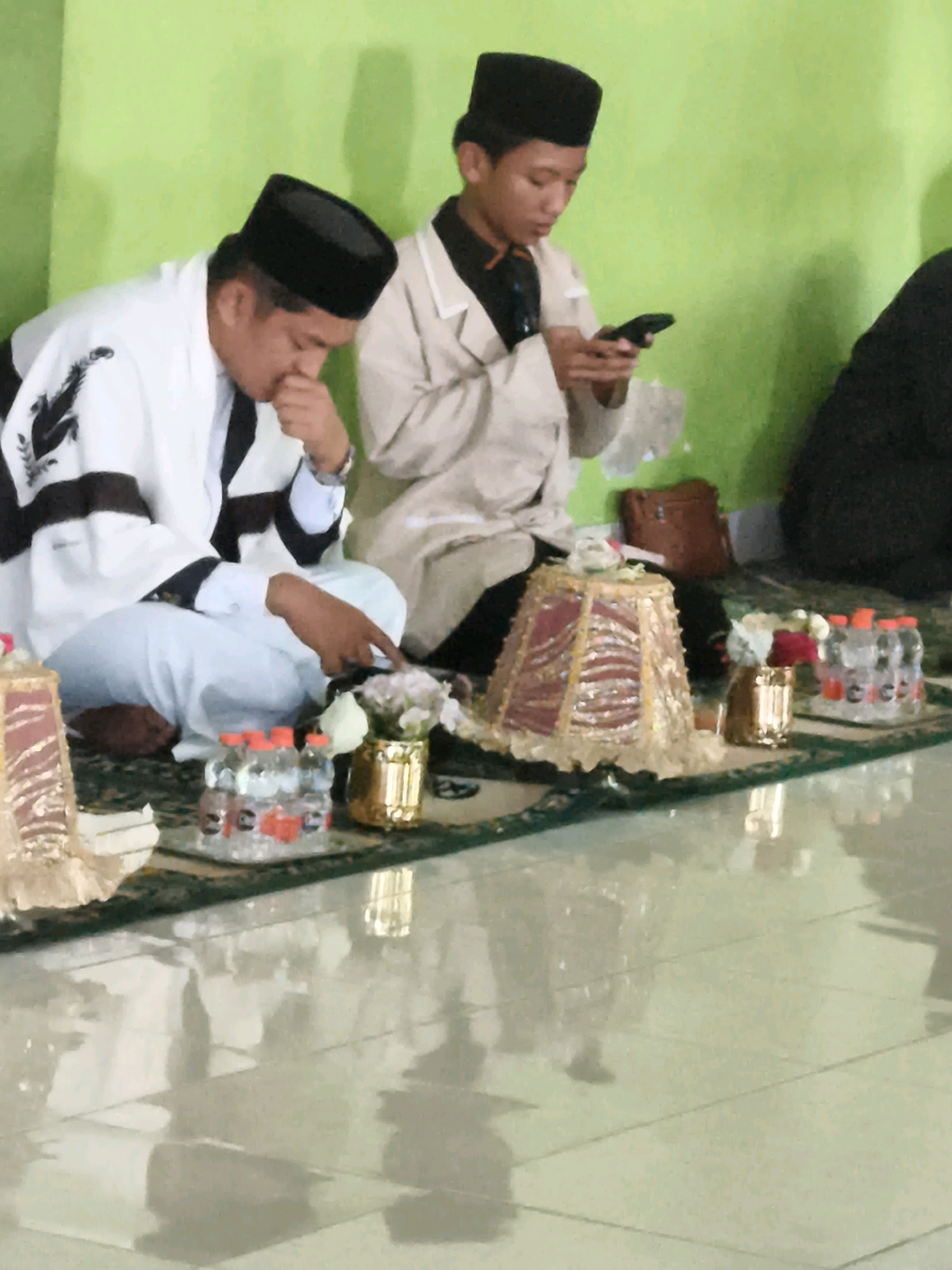 Two individuals, likely male, are seated on a green carpeted floor in what appears to be an indoor setting. The individual on the left is wearing a white robe with black trim and a black cap, and is looking down with his hand near his face. The individual on the right is wearing a light-colored outer garment over a dark shirt and a black cap, and is looking at and holding a smartphone. In front of them are several decorative objects and bottled water. The wall behind them is painted a bright green. The floor in the foreground is tiled and reflective, showing the reflection of the scene. There is a brown bag to the right of the individual on the right. The overall lighting suggests an indoor event or gathering. The image is slightly blurry and appears to be taken from a moderate distance. The scene is indicative of a cultural or religious event, potentially in Makassar, Indonesia, given the attire and setting.