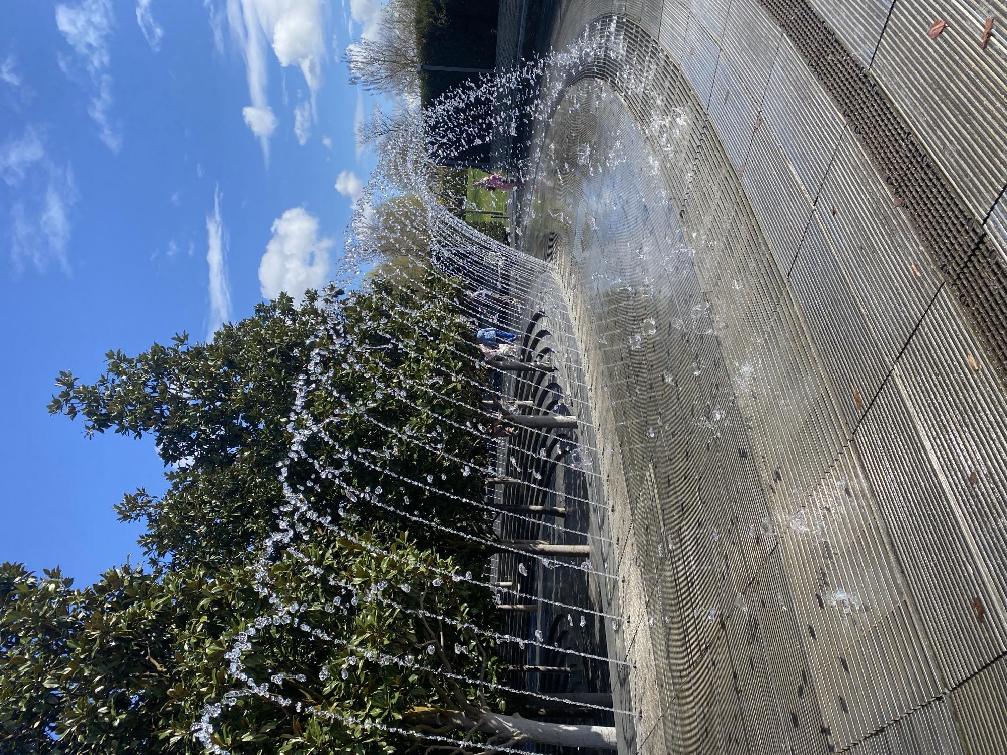 The image depicts an outdoor water fountain in the city of Krasnodar, Russia. The fountain is composed of multiple jets of water creating a curved archway. The fountain's base is made of gray, ridged concrete tiles. Lush green trees surround the fountain, partially obscuring a bright blue sky with scattered white clouds. In the background, figures of people are visible beyond the fountain, in a park-like setting with green grass. The lighting suggests a sunny day.