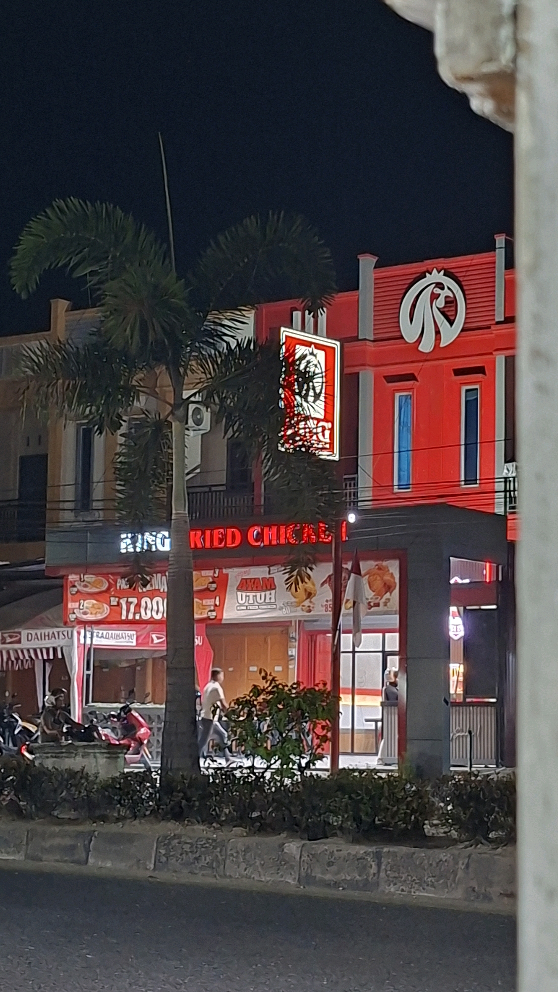 This is a nighttime, eye-level, medium shot of a KFC restaurant in Lhokseumawe, Indonesia. The building is predominantly red with white accents and features the KFC logo prominently. A palm tree stands in front of the building, partially obscuring a neon sign that reads "KING FRIED CHICKEN" in red lettering. Below this sign is a red banner displaying food items and prices, including an offer for "AYAM UTUH" (whole chicken). A white Daihatsu banner is visible on the left side of the image. Several people are present: a man in a white shirt walks past the building, another person sits on a bench with a motorcycle nearby, and a person stands inside the establishment. The street in front of the restaurant is visible, with a low concrete barrier and some green bushes. The overall scene depicts a bustling commercial area at night.