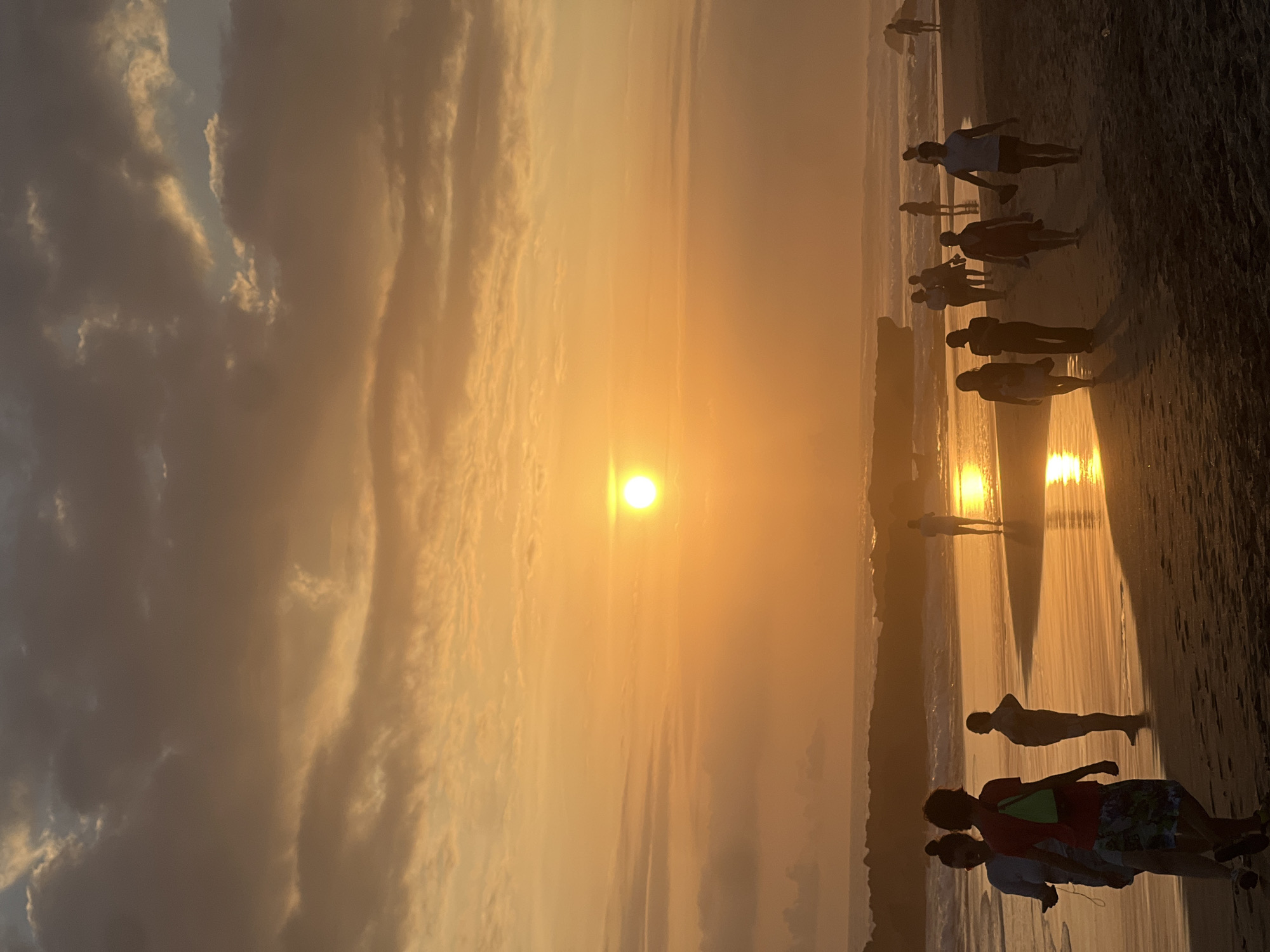 The image captures a sunset scene on a beach, likely in the North Kuta area of Indonesia, judging by the prevalent warm, golden light and tropical coastal setting. The sun, a bright, circular orb, is positioned in the upper left quadrant of the sky, casting a warm, orange-yellow glow that illuminates the clouds and the wet sand. The sky is a mix of soft oranges, yellows, and muted blues, with scattered clouds adding texture and depth.
A group of people are silhouetted against the vibrant sunset, walking along the shoreline. In the foreground, two individuals, possibly a young woman and a man, are walking towards the viewer, their figures dark against the luminous sand. The woman on the left appears to be holding a phone. To their right, a single person stands facing away from the viewer, their posture suggesting contemplation or observation of the sunset. Further down the beach, a larger group of people are scattered, some walking in pairs or small clusters, others appearing to be walking individually. They are all silhouetted, their features indistinguishable. The wet sand near the water reflects the warm hues of the sunset, creating a shimmering path.
The ocean is visible in the background, with gentle waves breaking on the shore. The water is also reflecting the sunset colors. Along the horizon, a rocky outcrop or a breakwater is visible, its dark form silhouetted against the bright sky. The overall atmosphere is serene and tranquil, typical of a beach at sunset. The lighting conditions are characteristic of the "golden hour," with soft, diffused light creating long shadows and a warm color palette. The image appears to be taken from a slightly low angle, emphasizing the vastness of the sky and the expanse of the beach.