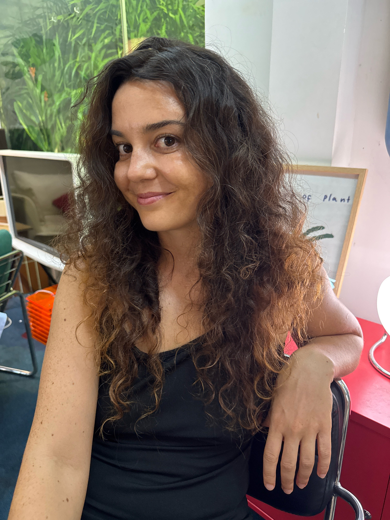 A young woman with long, curly brown hair and a black tank top is sitting in a chair, with her right arm resting on the back of the chair. She is smiling and looking at the camera. In the background, there is a whiteboard with writing on it and some plants. The location is the city of Phú Thủy, Vietnam.