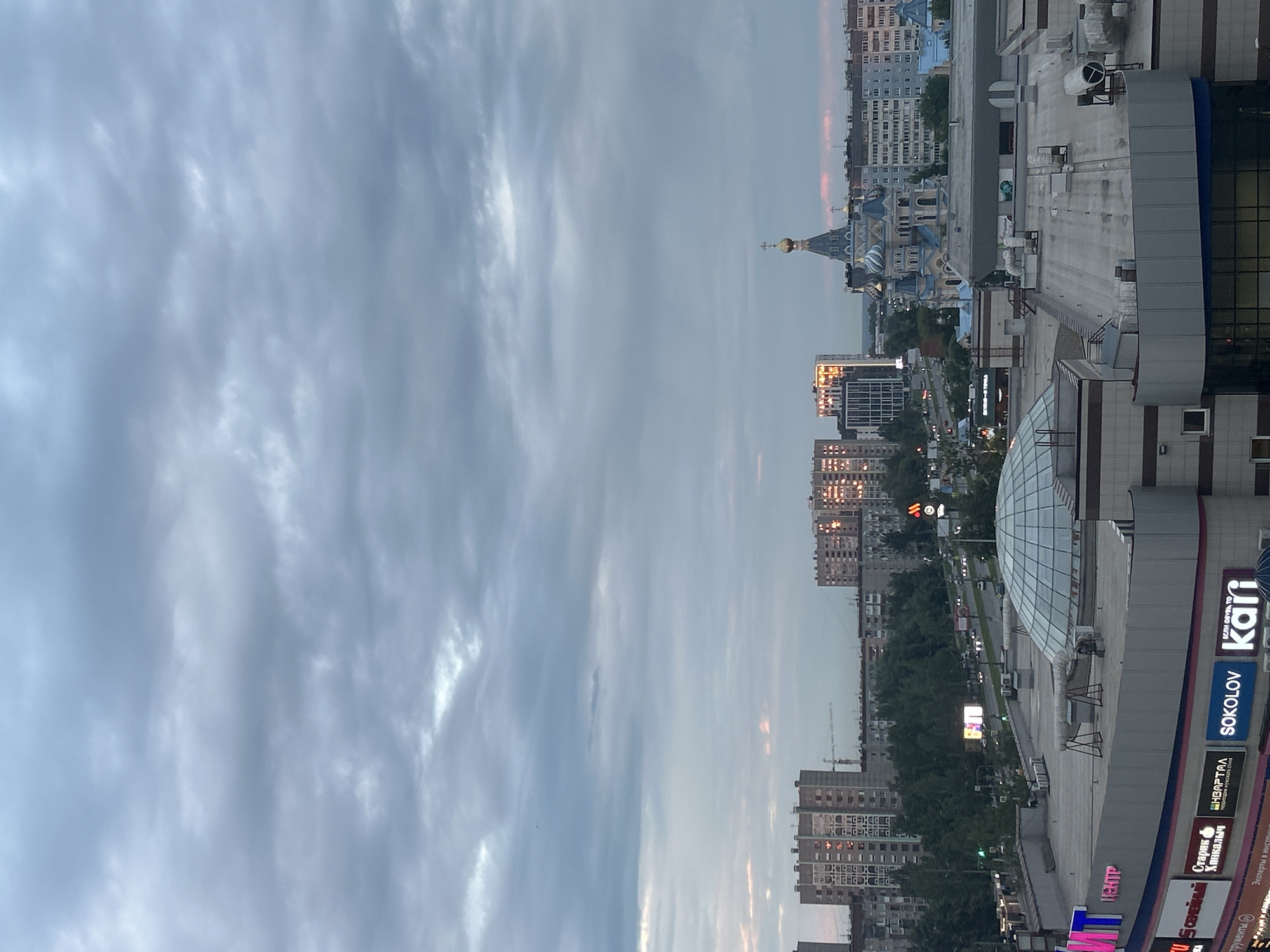 This is a wide-angle, eye-level, outdoor, daytime photograph of a cityscape, likely taken from an elevated position. The sky is overcast with thick, grey clouds. In the mid-ground, a large, multi-domed blue and white church with a golden cross on its tallest spire is prominent. To the left of the church, several modern buildings are visible, some with reflective windows that catch the light, suggesting the sun is setting or rising. A busy road with cars can be seen running through the city. In the foreground, the roof of a commercial building is visible, featuring a large glass dome structure and several air conditioning units. Several signs with Russian text are present on the building in the foreground, including "ЦЕНТР", "Sokolov", and "kari". This scene is characteristic of Izhevsk, Russia, likely depicting the area around the Holy Trinity Cathedral. There are no people clearly visible in the image.