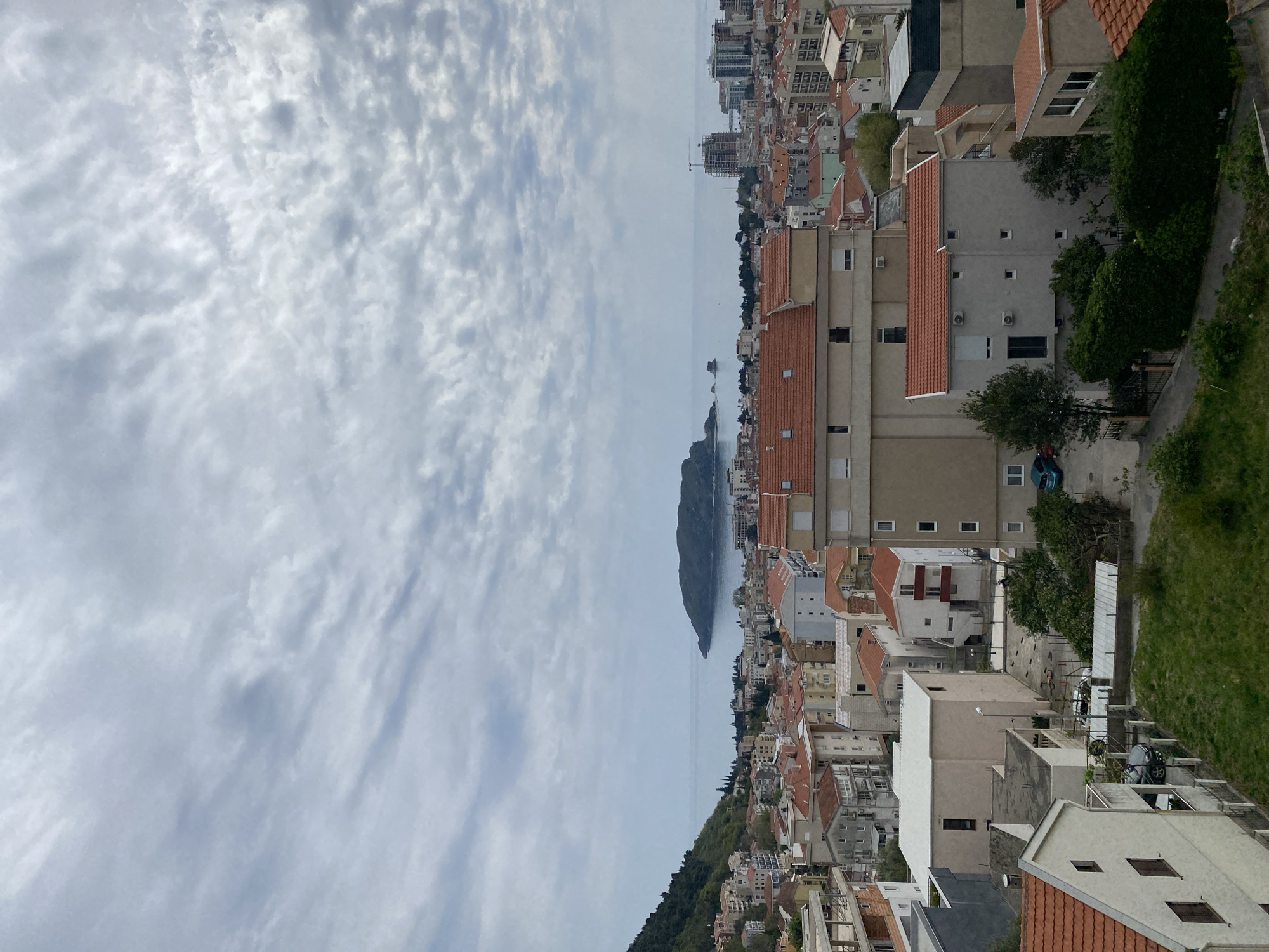 A landscape shot taken from an elevated vantage point in Budva, Montenegro, depicts an overview of the city. The scene includes various buildings with red-tiled roofs, interspersed with occasional patches of greenery and trees. A narrow stretch of water separates the urban area from a nearby island or peninsula. The sky above is filled with clouds, suggesting an overcast day. There are no visible people or actions occurring in the shot.