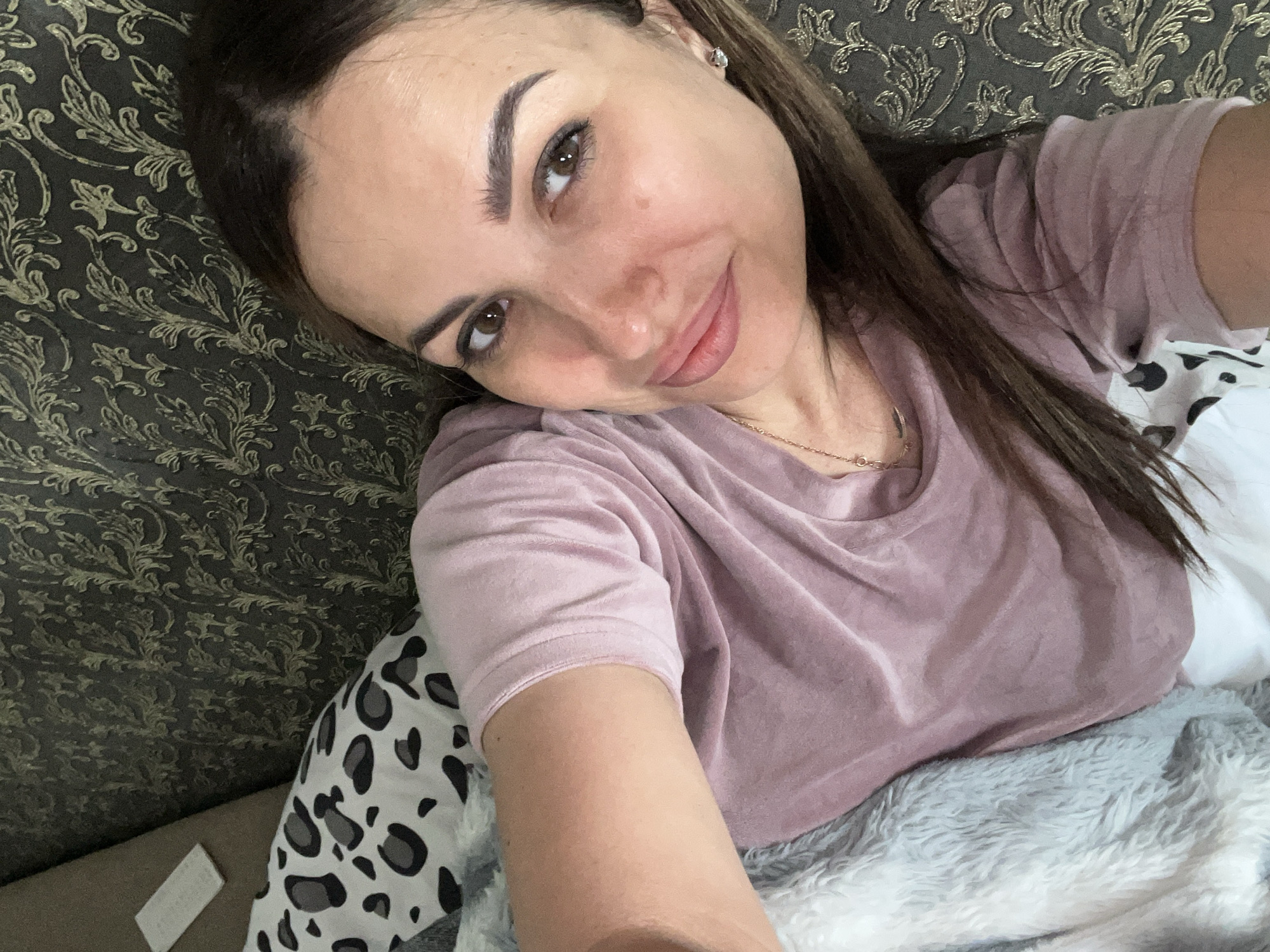 A woman with brown hair and brown eyes is lying down and looking at the camera. She is wearing a pink t-shirt. She has on makeup and a small silver earring in her right ear. She is lying on a couch with a patterned pillow and a fluffy gray blanket. The background is a dark couch with a gold floral pattern.