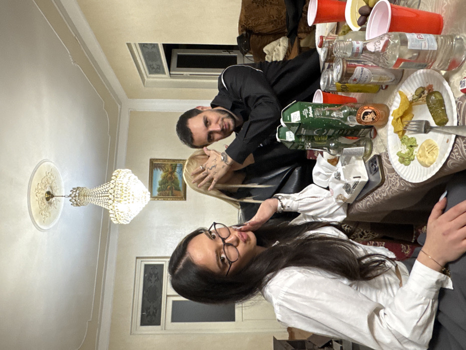 An indoor scene in Arinj, Armenia, shows three individuals seated around a table, engaged in a social gathering. The room features light-colored walls, a white ceiling with decorative molding, and a crystal chandelier. A framed landscape painting hangs on the wall, and two doorways or windows are visible in the background.
From left to right: A female with long dark hair, wearing glasses and a white puffy-sleeved blouse, is posing with her right hand near her cheek and her lips pursed towards the camera. Next to her, a female with blonde hair, wearing a dark jacket, has her face obscured by the hands of the male to her right. Multiple rings are visible on her fingers. The male, with dark hair and a beard, wearing a black collared shirt with a white stripe, looks towards the camera while holding his hands over the blonde female's face. Tattoos are present on the back of his right hand.
The table in the foreground is covered with a patterned tablecloth and holds various items. A white paper plate contains tortilla chips, two dips (one green, one light-colored), and a large pickle, with a fork resting on it. Several beverages are present, including green-boxed cartons, glass bottles (one clear labeled "PELLEGRINO," one amber labeled "Thomas Henry"), aluminum cans (one silver labeled "PELLEGRINO"), and multiple red plastic cups. A pack of tissues and a smartphone are also on the table. A brown upholstered sofa is partially visible on the far right.