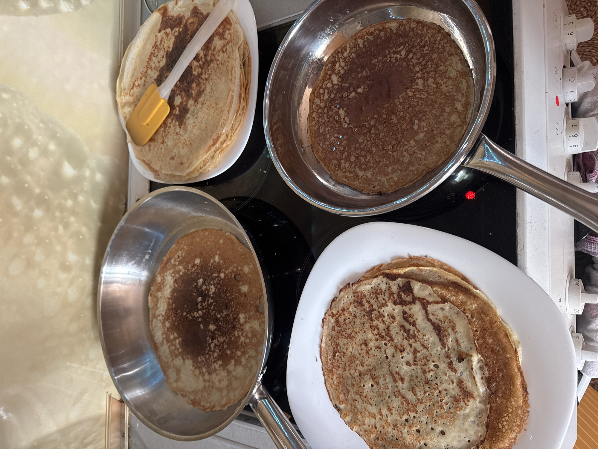 This overhead shot captures a domestic kitchen scene in Vysoky, Russia, centered around the activity of making pancakes or blini. Two metal frying pans sit on a black electric stovetop, each containing a thin, round pancake in different stages of cooking. One pancake in the top-right pan appears deeply browned, while the one in the bottom-left pan is lighter with distinct cooked patterns. Red indicator lights suggest that at least two burners are active.
Beside the stovetop, two white oval plates hold stacks of freshly cooked pancakes. The stack in the top-left features a yellow and white silicone spatula resting on it. The pancakes display varying shades of golden brown, with some exhibiting darker, crispier areas. To the right of the stovetop, white rotary control knobs for the appliance are visible, with numbers such as "0", "1", "2", "7", "8", "9", "50", and "300" discernible on them. A small red light and an 'S'-shaped icon are also near these controls. Part of a light-colored, textured surface, possibly a countertop, is visible on the far left, along with a glimpse of a textile on the far right. The scene depicts an ongoing or recently completed cooking session, likely for a meal such as breakfast or brunch.