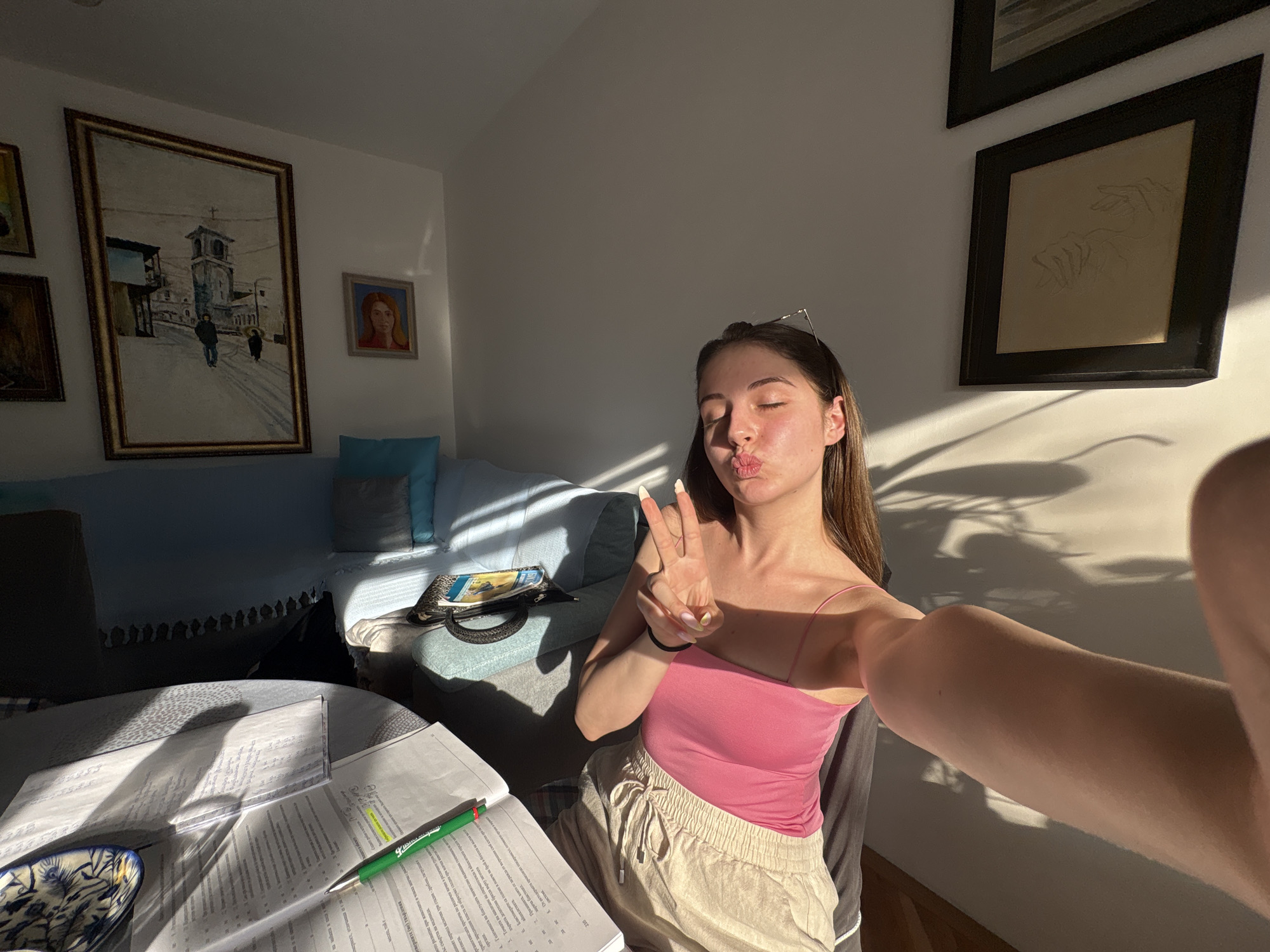 A young woman with long brown hair is taking a selfie in a brightly lit room. She is wearing a pink tank top and beige shorts. Her eyes are closed, and she is puckering her lips while holding up two fingers in a peace sign.
The room appears to be a living area or study. Sunlight streams in from a window, casting long shadows across the walls and furniture. On the wall behind her are several framed paintings. One large painting depicts a European town square with a bell tower, and another is a sketch of hands. To her left is a blue sofa with pillows, and in the foreground, a round table is covered with papers and a green pen. The papers have text in what appears to be Cyrillic script, and the OCR indicates some are scientific or academic notes. The lighting suggests it is either morning or late afternoon.