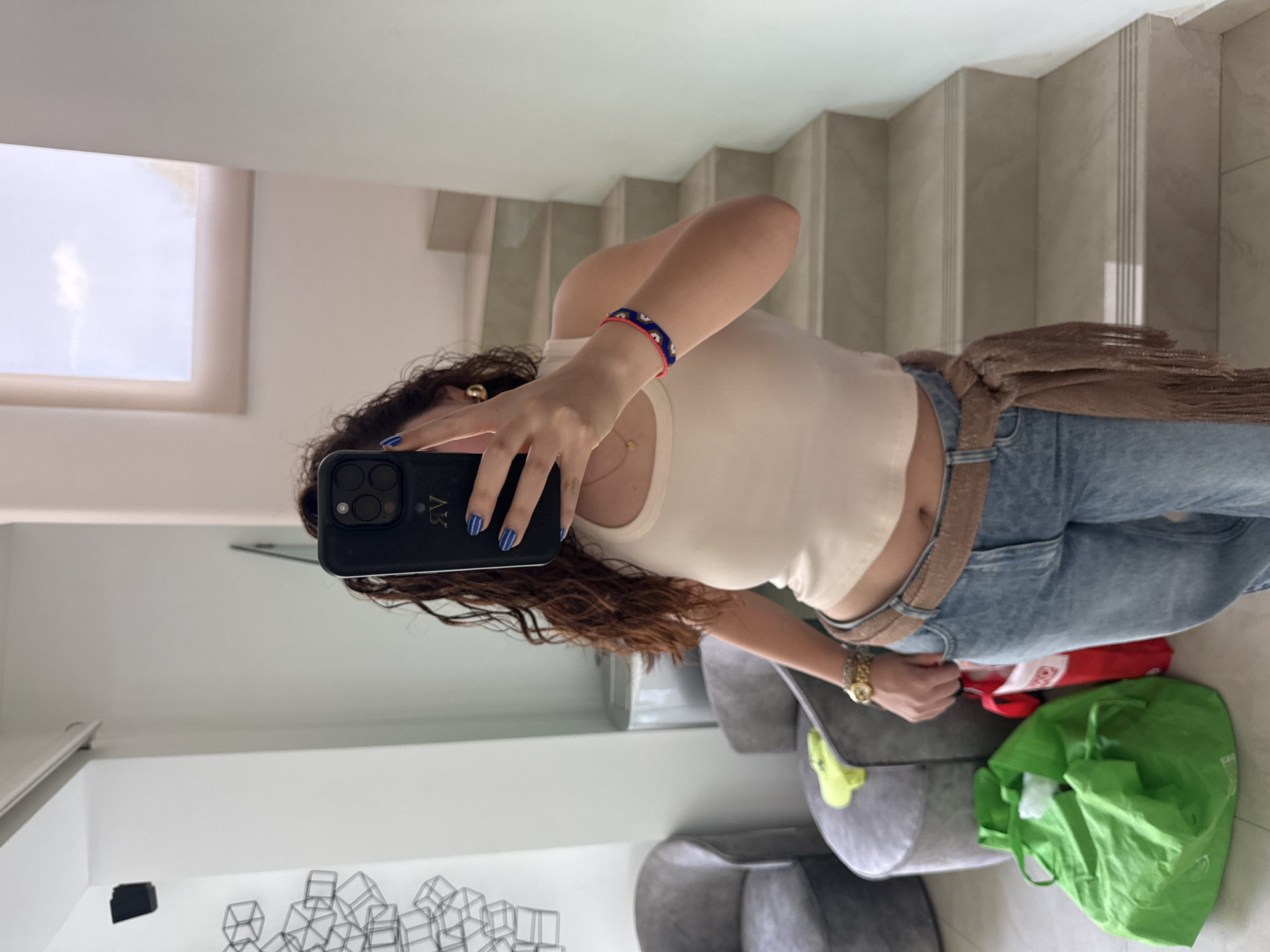 A woman is taking a selfie in a mirror. She is holding a black phone with a monogram in her right hand. She is wearing a beige top, blue jeans with a brown belt, and a bracelet. Her nails are painted blue with white stripes. The environment includes a staircase, a window, furniture, and shopping bags. A grid-like sculpture is on the wall. The image appears to be taken indoors, potentially in a home. There is no indication of Santa Clara Ocoyucan, Mexico.
