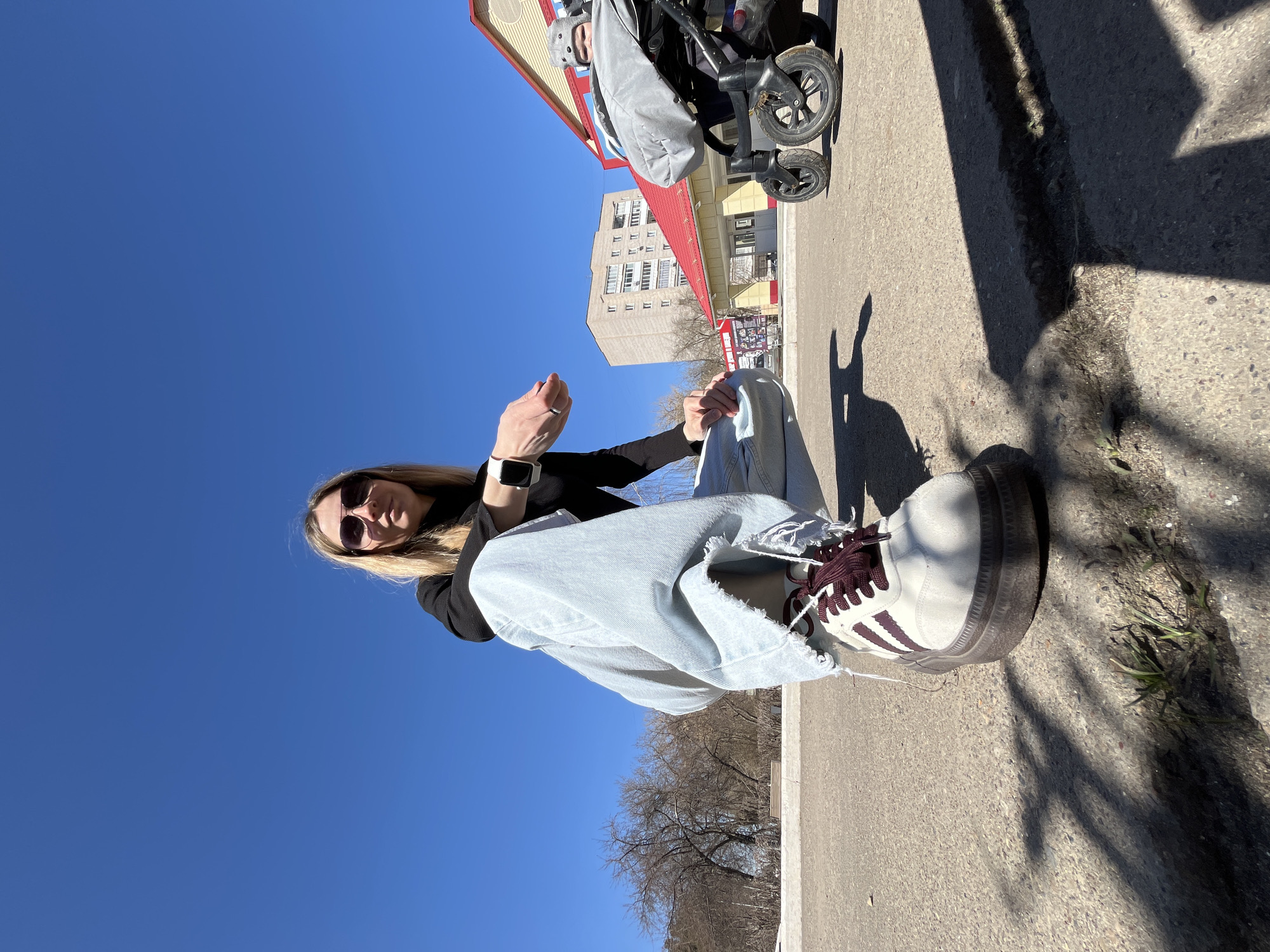 A young woman sits cross-legged on a sidewalk in Chaikovsky, Russia, facing the camera. She wears dark sunglasses, a black long-sleeved shirt, light blue ripped jeans, and white sneakers with burgundy stripes and laces. A smartwatch is visible on her left wrist. Behind her, trees line a low wall. In the background, buildings with red roofs are visible, along with a stroller. The sky is clear and blue. The sidewalk is made of concrete.