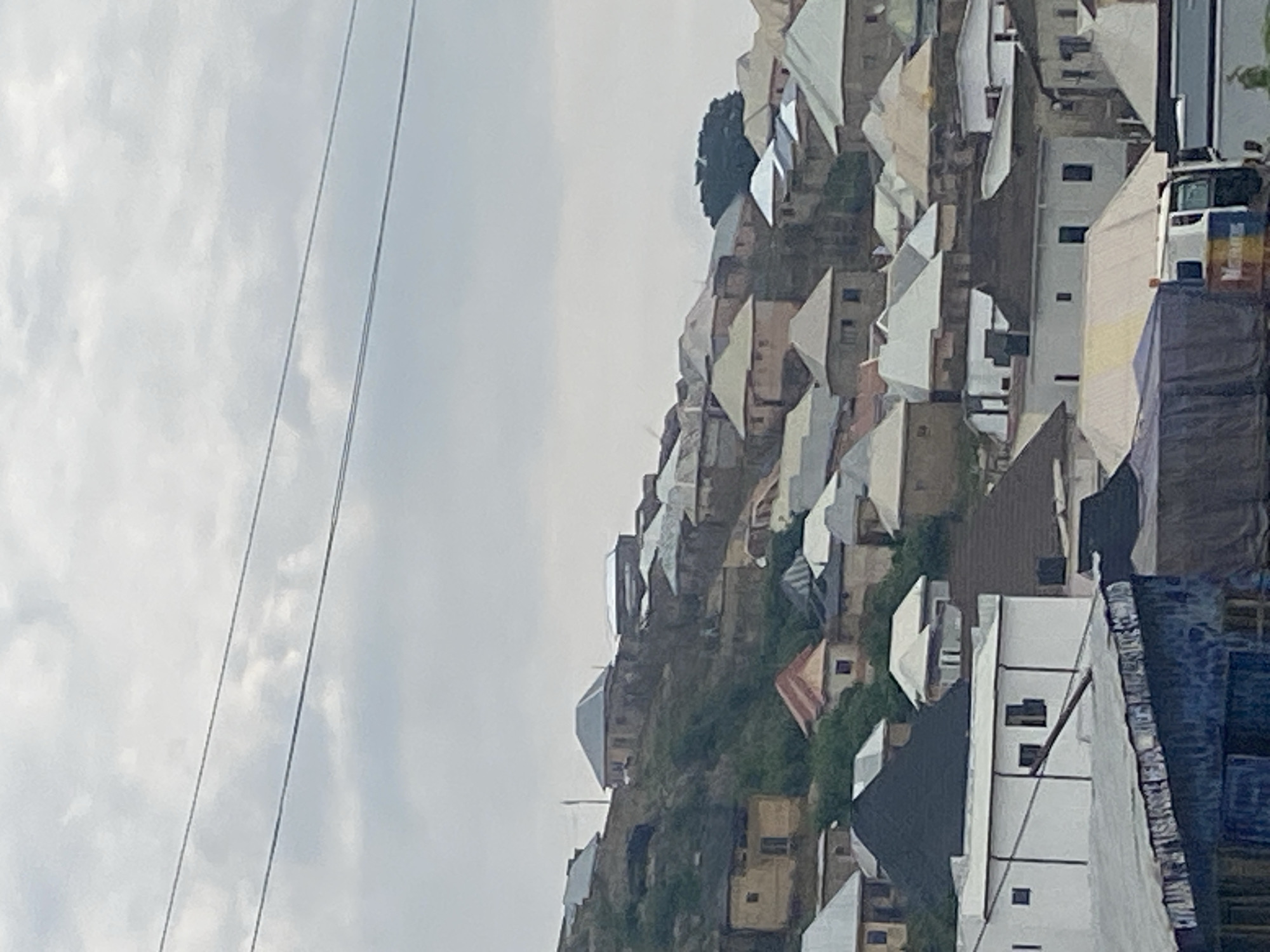 The image captures a hillside settlement in Jos, Nigeria, characterized by a dense collection of buildings with varied roofing materials. The architecture suggests a mix of traditional and modern influences, with many structures featuring corrugated metal or concrete roofs. The buildings are closely packed together, clinging to the natural contours of the hilly terrain. Vegetation is visible interspersed among the structures, indicating a semi-urban or peri-urban environment. The sky is overcast, casting a diffused light over the scene. In the foreground, parts of a large truck covered with a tarp are visible, along with other structures that appear to be part of a market or storage area. The overall impression is one of a vibrant community situated within a challenging, hilly landscape.