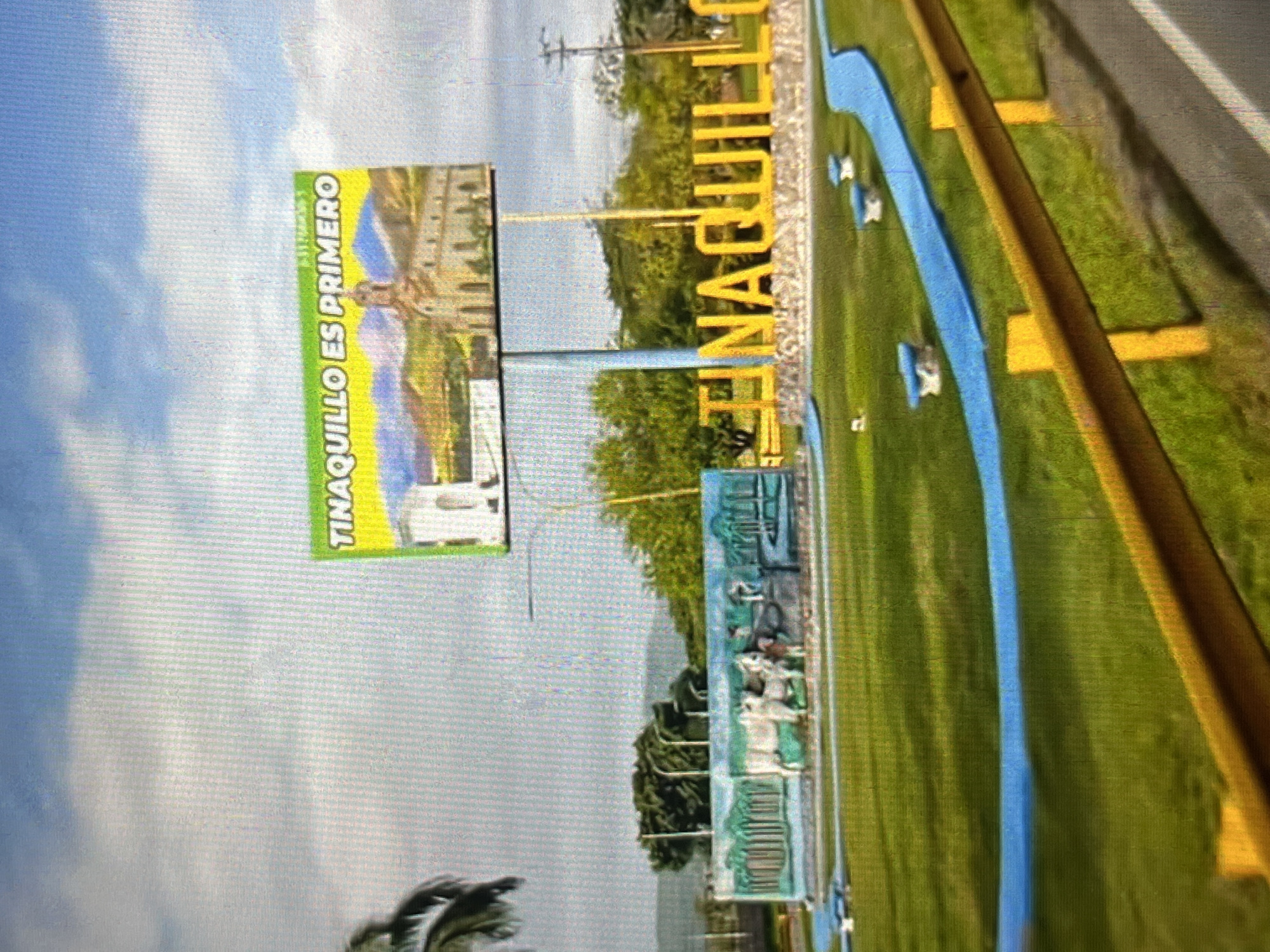 The image shows a roadside display in Tinaquillo, Venezuela. The display includes a billboard with the text "TINAQUILLO ES PRIMERO" (Tinaquillo is First) and an image of a building. Next to the billboard, there is a large sign spelling out "TINAQUILLO" in large yellow letters. The sign is surrounded by grass and a blue, river-like feature. A yellow barrier runs along the edge of the scene, likely adjacent to a road.