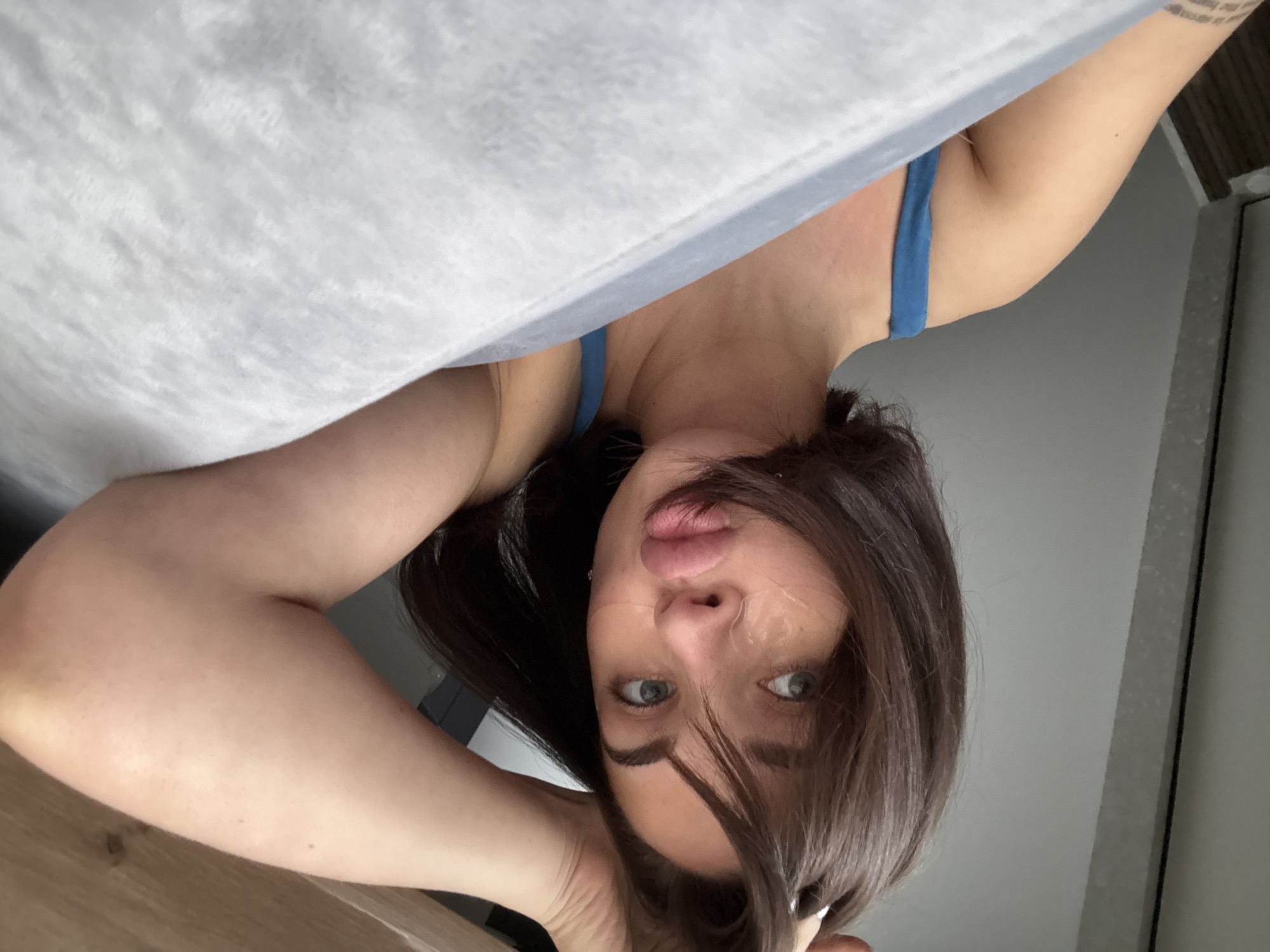 A woman with brown hair and blue eyes is lying in bed, looking at the camera. She has a blue tank top on and is partially covered by a light grey blanket. She appears to be in an indoor environment. There is no indication of the city of Лобаново, Russia.