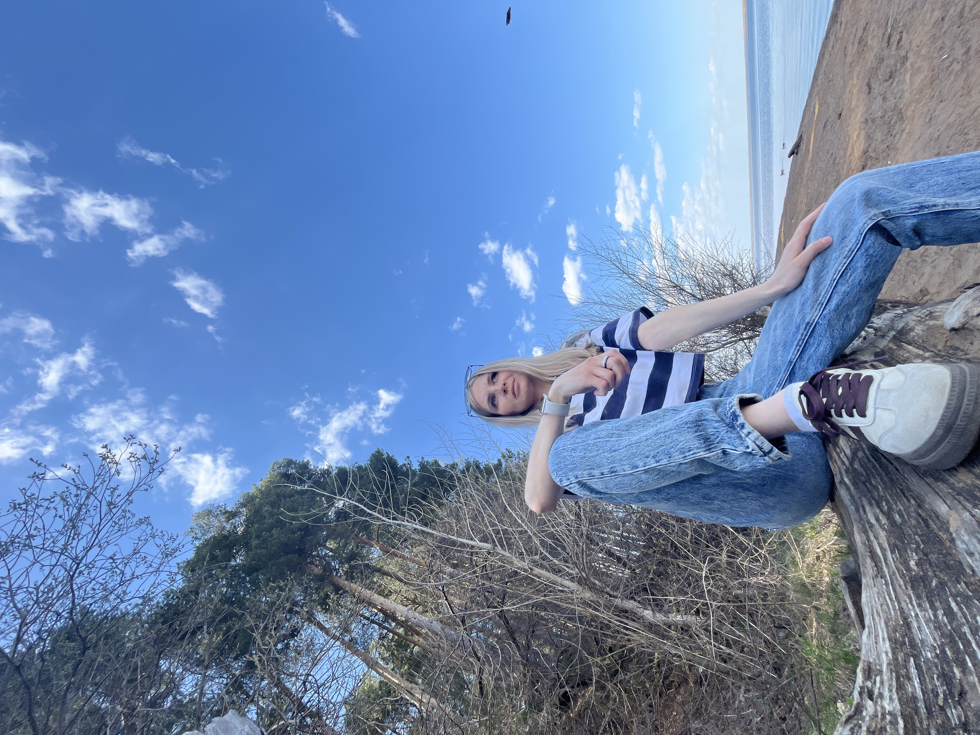 A young woman is sitting on a fallen tree trunk in Chaikovsky, Russia. She is wearing jeans, a striped top, and sneakers. The location appears to be near a body of water (presumably the Kama River or a smaller lake), and there are trees in the background. The sky is blue with some clouds. The photo is taken from a low angle, looking up at the woman and the sky. A bird is visible in the top center of the frame.
