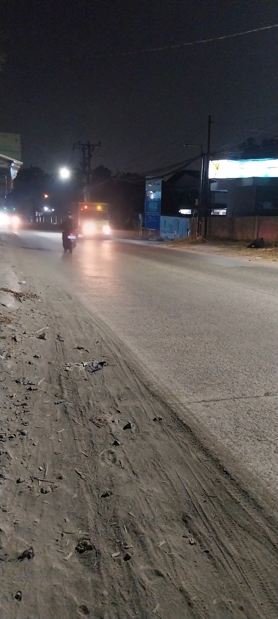 The image captures a night scene on a road in Kelapa Dua, Indonesia. In the foreground, the sandy and dirt shoulder of the road is visible, with debris and tire tracks. The road itself is paved and illuminated by the headlights of a vehicle, appearing wet or reflective. A motorcycle is seen in the middle ground, also illuminated by its headlights. Further down the road, a larger vehicle, possibly a bus, with its headlights and taillights illuminated, is approaching. Buildings and a utility pole with wires are present on the right side of the road, with signage indicating "PLN". The overall environment suggests a moderately urban or suburban area at night.