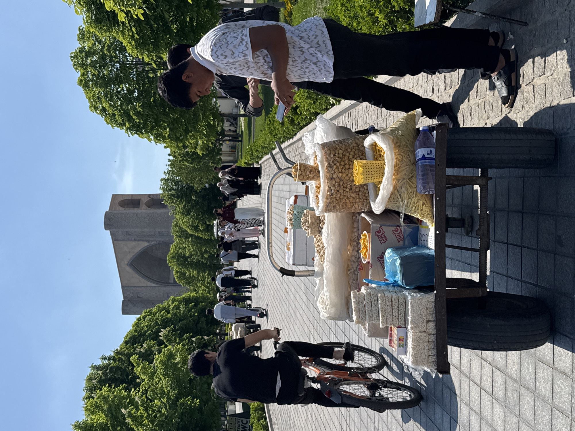 A street scene in Samarkand City, Uzbekistan, shows two men near a cart filled with snacks, nuts, and bottled water. One man is on a bicycle. The other is standing and looking at his phone. A group of people walks along a path behind the cart. A large, ornate building is visible in the background, surrounded by green trees under a clear, bright sky. The ground is paved with grey stone tiles.