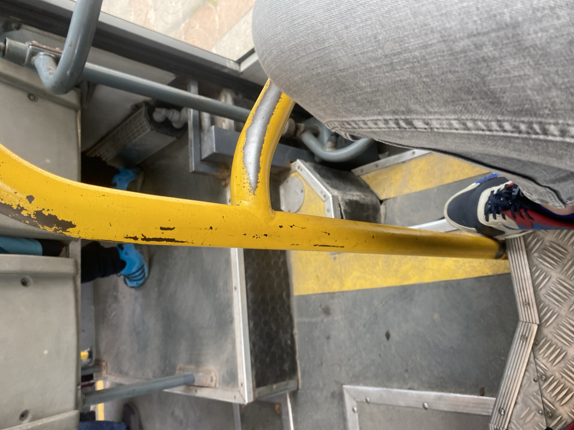The image depicts the interior of a public transport vehicle, likely a bus, as evidenced by the presence of a yellow handrail, metal flooring with grip texture, and seating structures. The perspective is from a seated passenger looking downwards and slightly forward.
Dominating the foreground is the lower portion of the passenger's right leg, clad in light-wash denim jeans. The passenger is wearing a sneaker with a predominantly navy blue and white design, accented with red stripes and black laces. Adjacent to the passenger's leg is a substantial yellow metal pole, a common feature in public transport for stability. This pole exhibits signs of wear, with chipped yellow paint revealing a metallic grey underneath in several areas.
The flooring of the bus is a mosaic of materials. Directly beneath the passenger's feet is a diamond-patterned metal plate, likely for enhanced grip. This transitions into a grey, textured surface with a distinct yellow strip running diagonally across it. Further back, a dark, textured material is visible, possibly a mat or a specialized section of the floor.
In the mid-ground, the legs of other passengers are visible. One individual is wearing bright blue athletic shoes with white stripes, characteristic of a popular sports brand. They are also wearing dark trousers. Another passenger, partially obscured, is wearing dark footwear.
The environment suggests an urban setting, given the context of public transportation. Specifically, the image is located in Smolyan, Bulgaria, a city known for its mountainous terrain and historical significance, though these specific details are not visually evident from the image itself. The interior of the bus appears functional and utilitarian, with the yellow handrail serving as a focal point for safety and accessibility within the vehicle. The lighting suggests it is daytime, with ambient light entering from an unseen window.