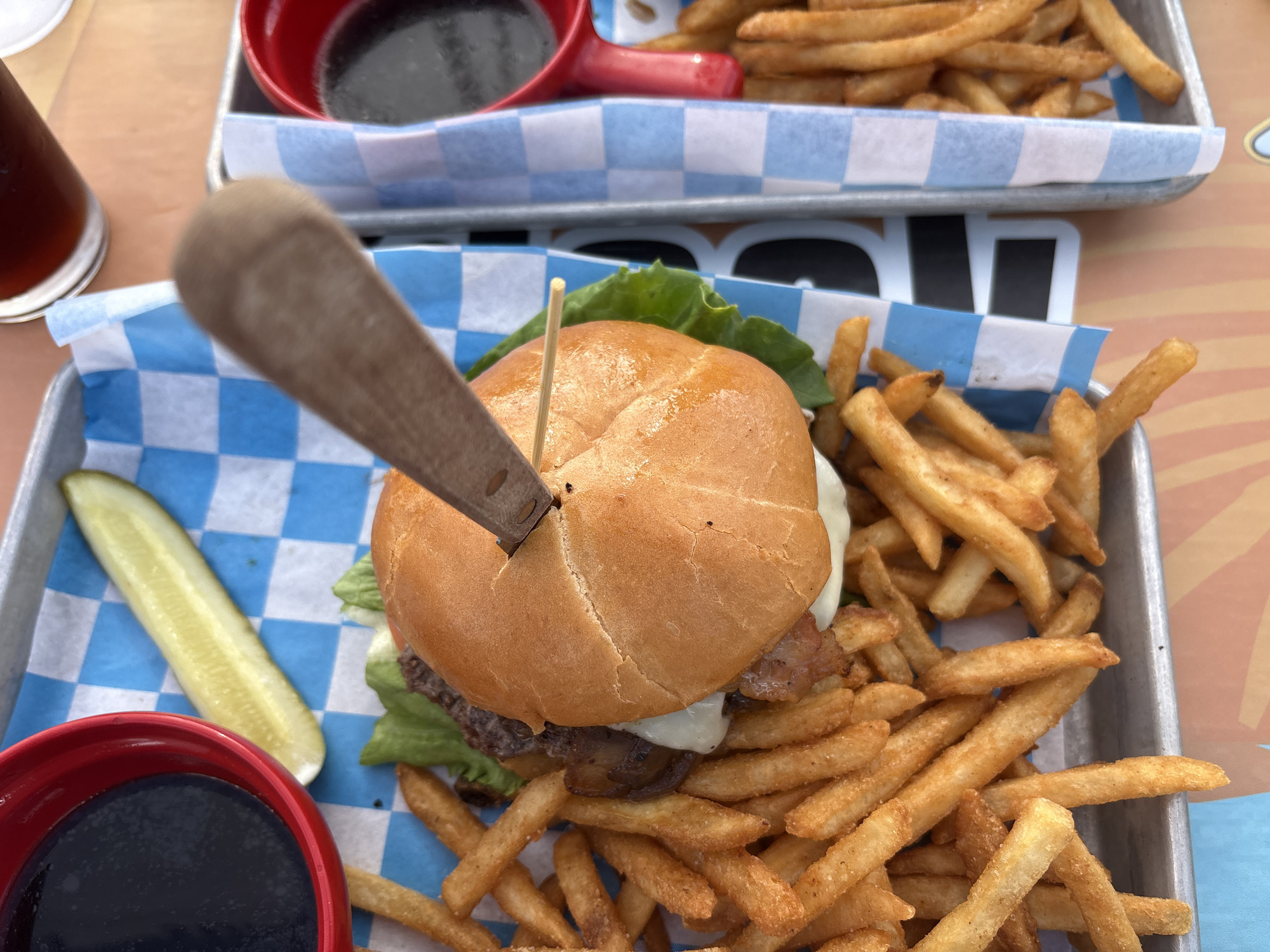 The image shows a close-up, overhead view of two metal trays filled with french fries and burgers, likely at a restaurant. The tray in the foreground contains a burger with a wooden skewer holding it together, a dill pickle spear, and a red dipping sauce bowl filled with a dark liquid. The burger is topped with lettuce and appears to have cheese and bacon. The second tray, partially visible on the right, also holds french fries and a red dipping sauce bowl. Both trays are lined with blue and white checkered paper. The scene is set on a surface with a striped pattern. The image appears to have been taken in Portland, Oregon, United States of America, judging by the distinctive checkered paper and the overall aesthetic often found in casual dining establishments in the city. There are no people visible in the image.