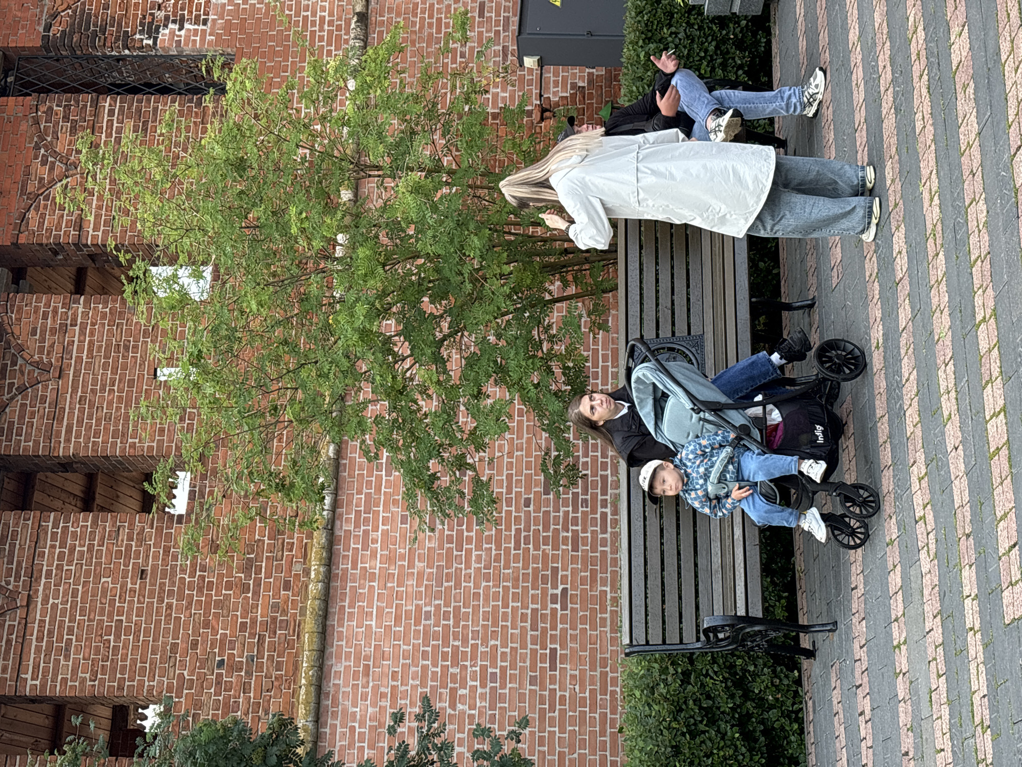 In Nizhny Novgorod, Russia, on a paved walkway in front of a red brick building, a group of people are gathered. A woman in a black jacket and jeans is seated on a wooden bench, looking to her left. Beside her, a toddler in a blue patterned jacket and jeans sits in a stroller. To the right of the bench, a person wearing a light-colored trench coat and jeans stands facing away from the camera, holding a phone. Another person, wearing a dark jacket and jeans, is seated to the far right of the bench, with a cigarette in their right hand. Green bushes line the area behind the bench, and a tree with lush green foliage stands behind the seated individuals. The brick building features arched openings and a metal-grated window.