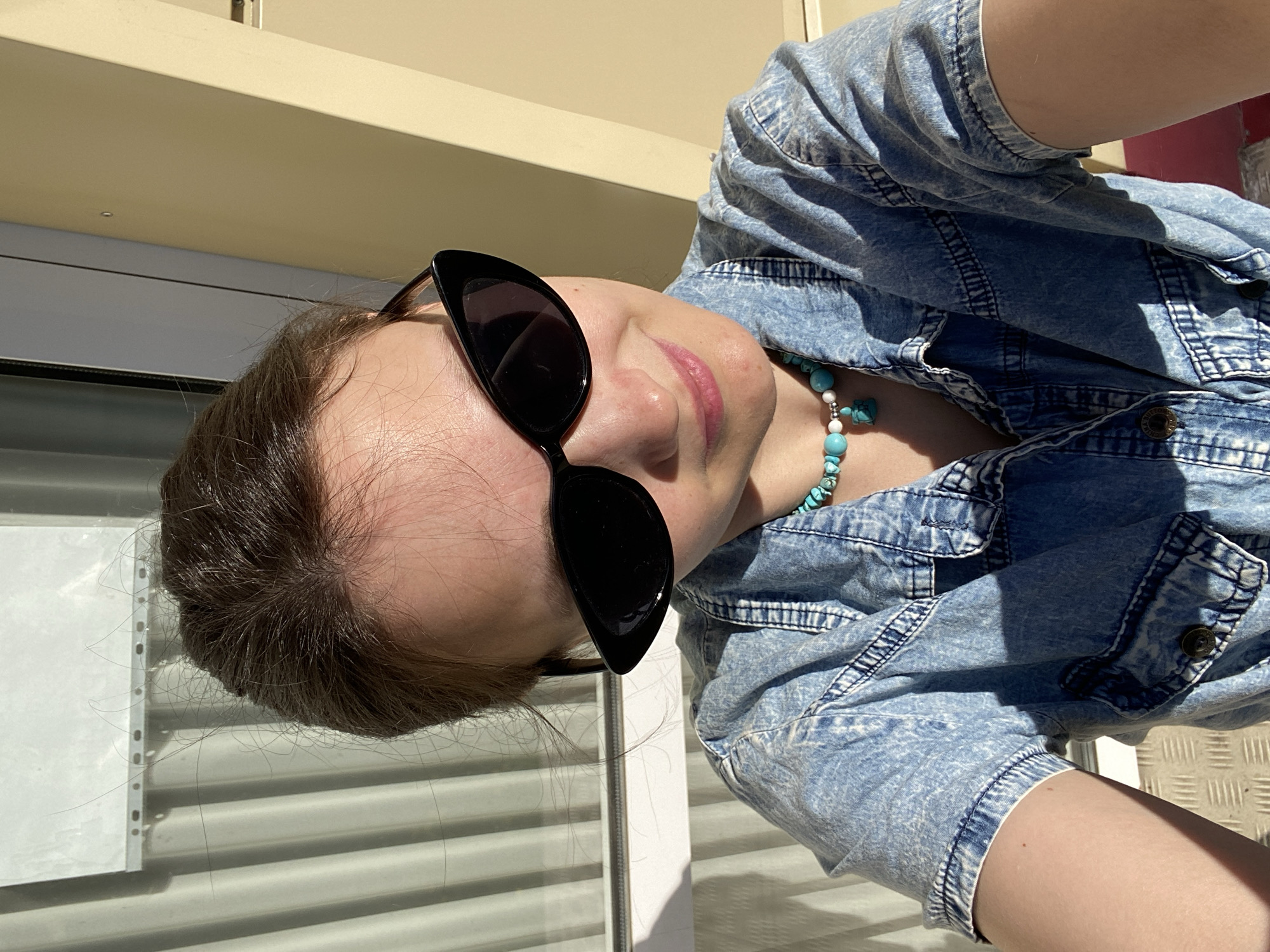 A woman wearing sunglasses and a light blue denim shirt is the subject of the image. She is also wearing a turquoise necklace. The background includes a white wall, a set of rolling blinds, and a tan or beige overhang. The image appears to be taken outdoors in bright sunlight. The location is Chaikovsky, Russia.