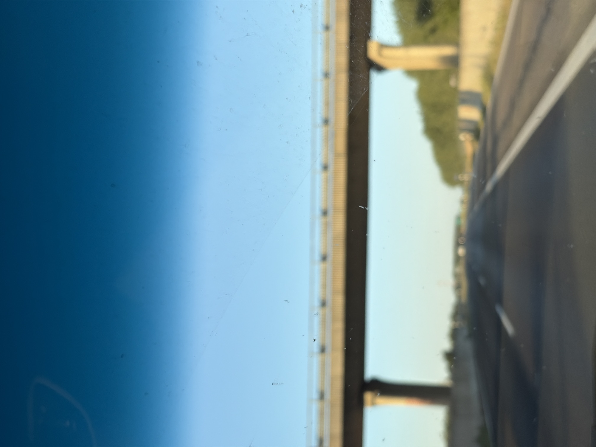 The image shows a view from a vehicle traveling on a highway. A concrete overpass with a metal railing runs horizontally across the upper middle portion of the frame. Below the overpass, the highway stretches into the distance, with a white lane marker visible on the right side. Trees form a line along the right side of the highway in the background. The sky is a clear blue, with a gradient of darker blue at the top of the frame. The overall scene suggests a sunny day. The image is out of focus and has numerous specks of dirt or dust on the surface it was captured through, likely a windshield. There is no discernible information regarding the city of Báb, Slovakia, within the visual content of the image itself.