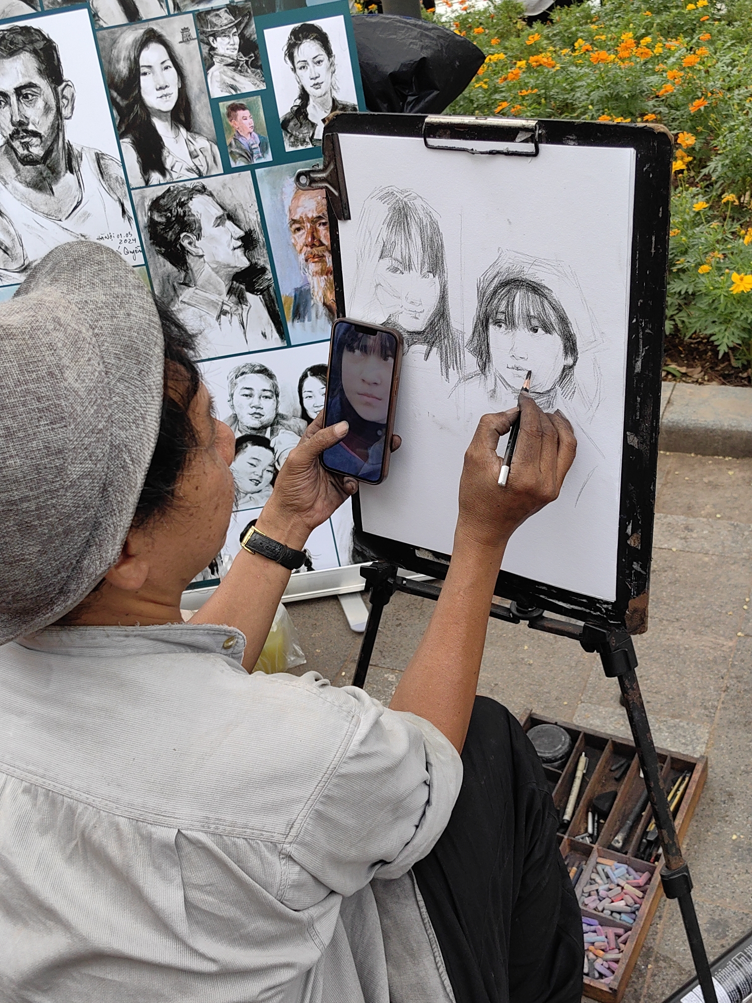 An outdoor portrait artist, viewed from behind, is depicted in the midst of creating new artwork. The artist, wearing a light gray shirt and a matching flat cap, holds a smartphone in their left hand, displaying a close-up portrait of a young girl as a reference image. With their right hand, they are actively sketching two female figures on a white sheet of paper clipped to an easel. The figures are rendered in pencil, with one being more detailed, featuring bangs and a youthful face, while the other is in an earlier, looser sketching stage.
Behind the artist, several finished portraits are displayed on a large board, showcasing a range of subjects and styles. These include a strong-jawed man with a mustache, a woman with long dark hair, a man in a hat, another woman with an intense gaze, and a child's portrait. An older man with a prominent beard and serene expression is also visible, possibly incorporating elements of color. One of the finished portraits clearly bears the handwritten text "Hà Nội 01.05.2024" and a signature that appears to read "Quyên".
At the artist's feet, a wooden box filled with various art supplies, including a vibrant assortment of pastels, pencils, and other tools, indicates a prepared and mobile workspace. The setting is outdoors, suggested by the paved ground and a lush bed of green plants with bright orange-yellow flowers in the background, indicating a daytime scene. The artist's focused posture and precise grip on the pencil convey concentration on their craft.