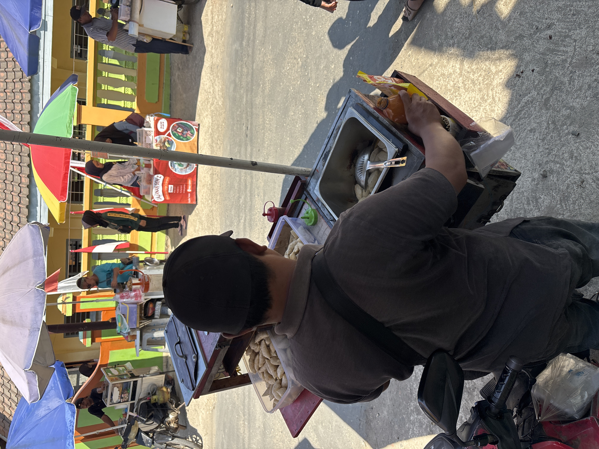 The image depicts an outdoor street scene in Tirtomoyo, Indonesia, characterized by a bright, sunny day casting strong shadows. In the foreground, a man wearing a dark t-shirt and a black baseball cap is seen from behind, standing next to a food cart. The cart is equipped with a metal sink containing a slotted ladle and what appear to be fried snacks, and a bin holding numerous small, oval-shaped food items. A bottle of orange sauce and a packet of snacks are also visible on the cart. The cart is mounted on a red base and appears to be attached to a motorcycle, indicated by the presence of a side mirror and handlebars in the lower left corner.
In the middle ground, several other food stalls are set up under colorful umbrellas. One stall features a red banner with text and images of food. People are visible interacting at these stalls, including individuals preparing or purchasing food. The background shows a building with a tiled roof and a yellow facade, behind a low wall adorned with green and orange accents. An Indonesian flag is also visible. The overall impression is a bustling street market or food vendor area.