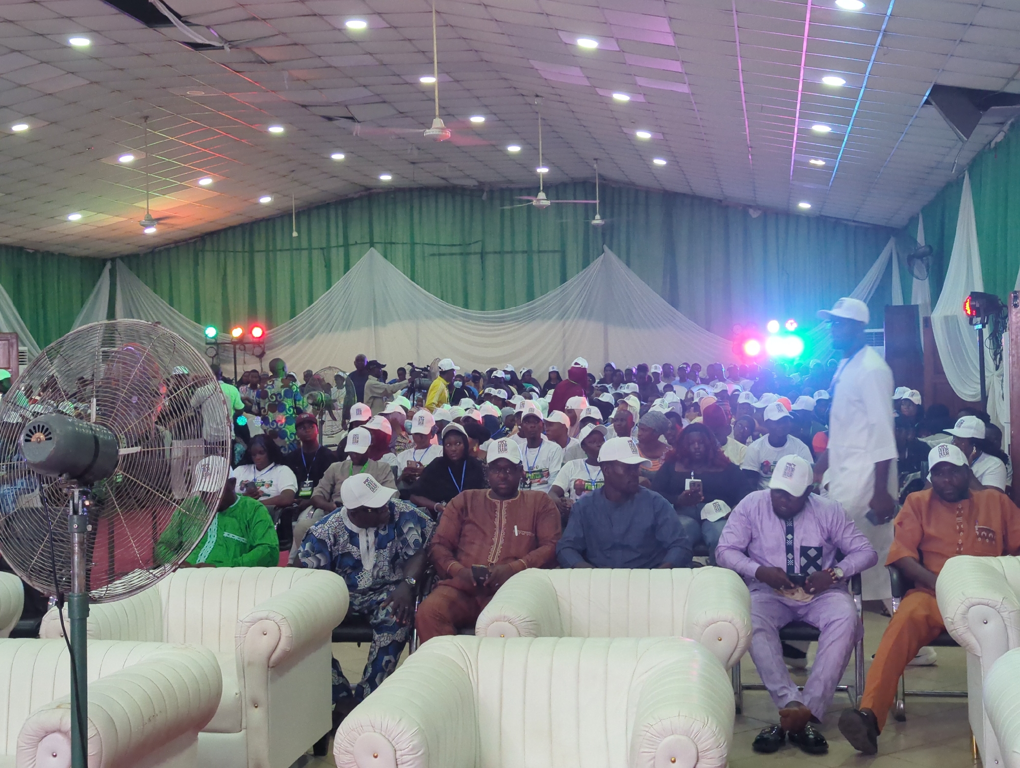 A medium, eye-level shot depicts a densely populated indoor event, likely a political gathering, in an unidentified location in Unknown, Unknown. In the foreground, there are multiple plush white sofas with individuals sitting on them. The people wear formal attire in various colors, many with white caps bearing what appears to be a logo or initial.
Beyond the foreground, a crowd of people fills the space. They wear the same style of white cap. The background features green and white draped fabric, suggestive of a tent or large indoor venue. Overhead lighting is profuse, with ceiling fans and multicolored spotlights illuminating the scene. A large oscillating fan is positioned prominently in the foreground.