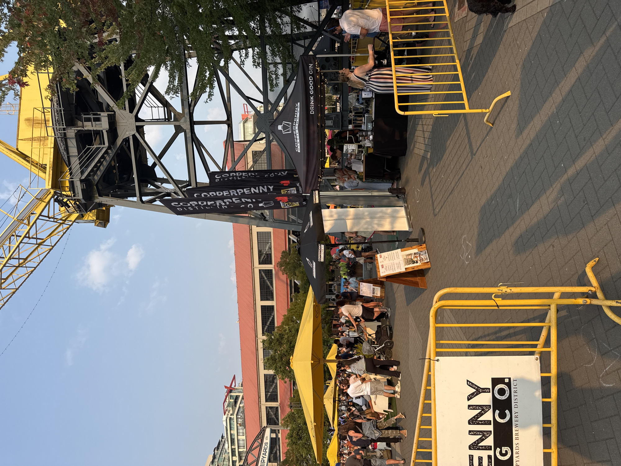 The image captures an outdoor event with a large yellow crane structure dominating the background. The location is identified as the Shipyards in the city of North Vancouver, Canada, as indicated by a sign that reads "SHIPYARDS LTD.". In the foreground, a yellow metal barrier encloses an area with a white sign displaying "GINNY G.CO." and "SHIPYARDS BREWERY DISTRICT".
Numerous people are visible milling about. Several yellow and black umbrellas provide shade over tables where people are gathered. A black tent with "COPPERPENNY DISTILLING CO." and "DRINK GOOD GIN" emblazoned on it is set up, with staff serving patrons. One woman in a black and white striped dress stands near the tent, interacting with someone. Other individuals are seen walking, standing, and sitting. The ground is paved with interlocking bricks, and long shadows suggest it is late afternoon or early evening. Trees are present, and modern buildings with red roofs are visible in the mid-ground. The sky is clear with a few scattered clouds.