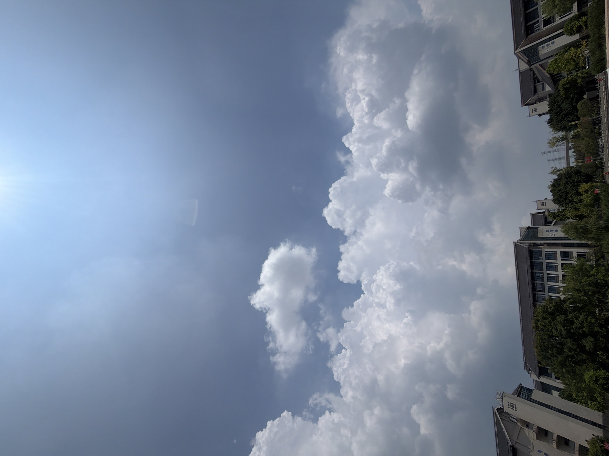 A wide-angle shot captures a clear blue sky with a sun flare at the top right. Scattered white cumulus clouds fill the upper half of the frame. Below the clouds, the tops of several buildings are visible, featuring dark tiled roofs and light-colored walls. Trees with lush green foliage are interspersed between the buildings. The scene appears to be an outdoor setting in a city, likely during the daytime with fair weather. The image's composition emphasizes the vastness of the sky.
