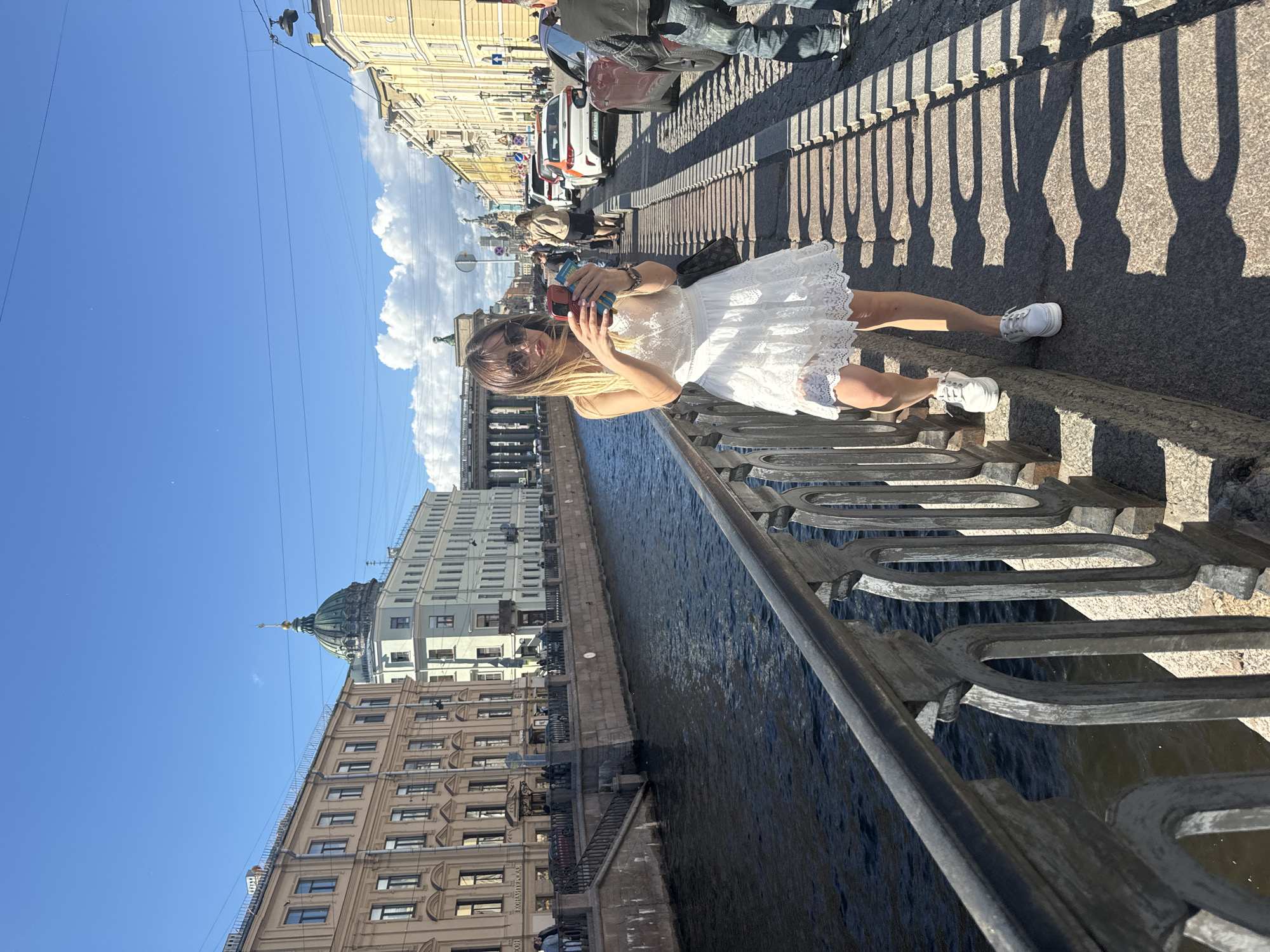 A young woman with blonde hair, sunglasses, and a white lace dress is standing on a bridge railing, holding a phone. She is on a sidewalk next to a canal in Saint Petersburg, Russia. Buildings with classical architecture line the canal. The sky is blue with some clouds, and there are cars parked on the street. The woman is looking at her phone and appears to be taking a picture or video. The overall mood of the image is bright and sunny, suggesting a pleasant day.