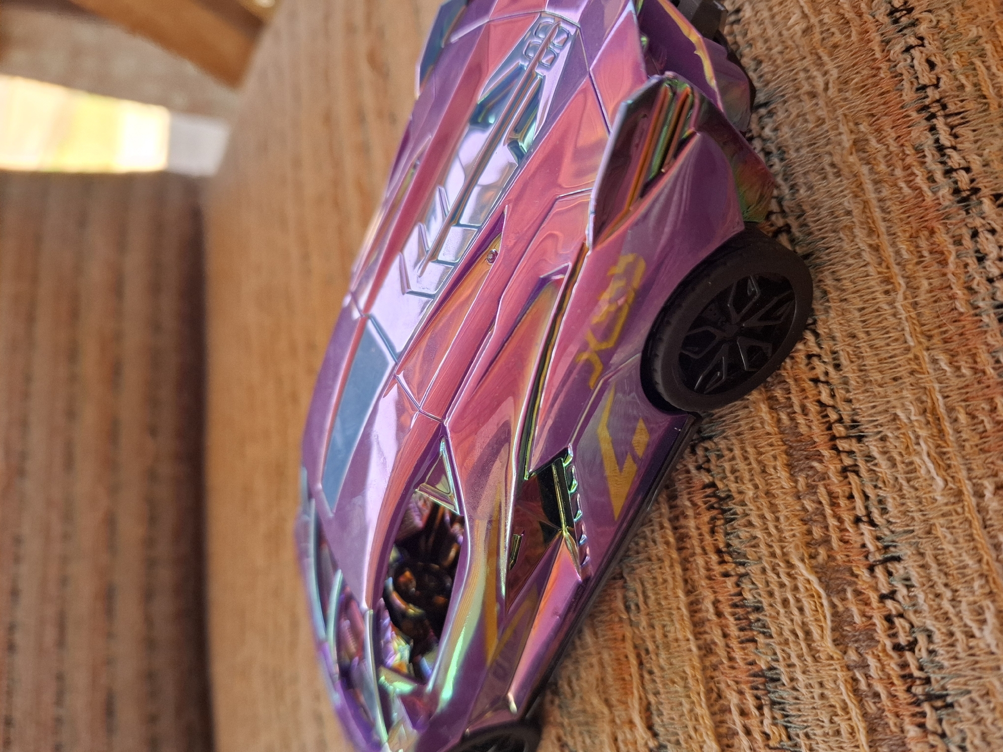 A close-up, low-angle, eye-level shot shows a detailed toy model of a futuristic sports car with an iridescent purple and pink finish, parked on a textured, woven surface. The car, with its sharp angles and sleek design, reflects the ambient light, creating a spectrum of colors. The interior of the car is visible, with black seats and a steering wheel. The background is blurred, suggesting a soft focus effect on the car. The scene appears to be indoors, with warm lighting. There is no identifiable context suggesting a location in Galabovo, Bulgaria.
