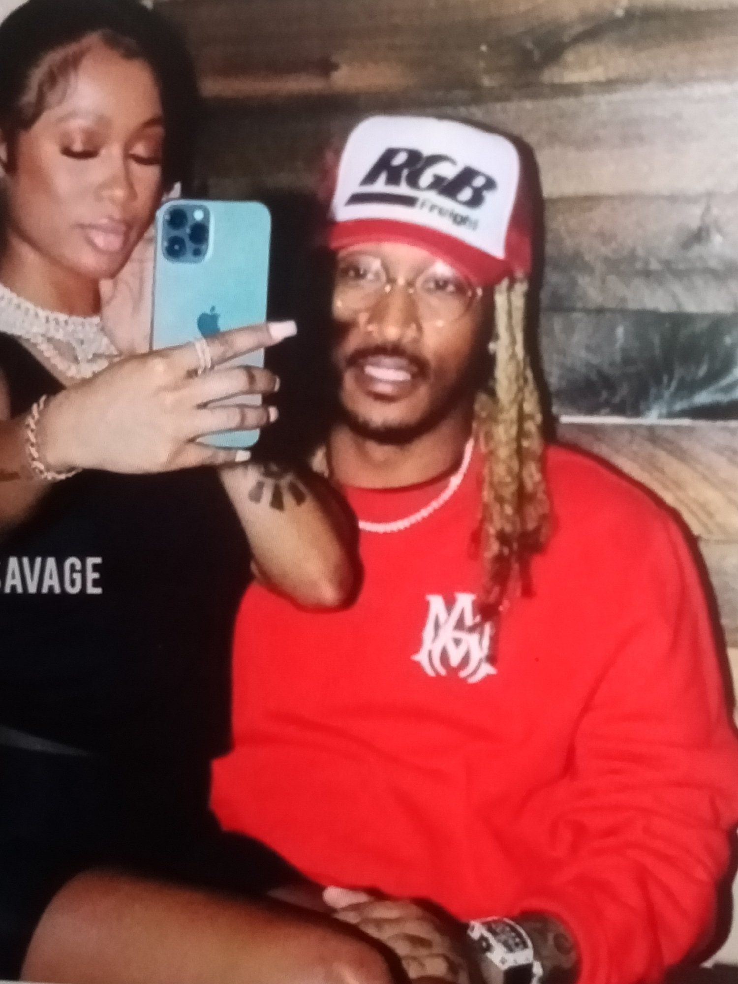 This is a waist-up, medium shot taken indoors, possibly in a restaurant or lounge with wood-paneled walls, showing two individuals. The person on the left, a woman with dark hair and makeup, is holding a light blue iPhone in her left hand, taking a selfie. She is wearing a black top with the word "SAVAGE" in white letters and a thick, diamond-encrusted necklace. A thin bracelet adorns her left wrist. The person on the right, a man with blonde dreadlocks and glasses, is wearing a red long-sleeved shirt with a white logo on the chest and a red and white trucker hat with "RGB Freight" printed on it. He is wearing a watch with a white band and black face on his left wrist. Both individuals appear to be looking towards the camera, and the woman's left arm is around the man's shoulder. There is a tattoo on the woman's visible arm and multiple tattoos on the man's visible arm. The lighting suggests it is an indoor setting, possibly at night. The mention of Jalingo, Nigeria, is not supported by any visual evidence within the image.