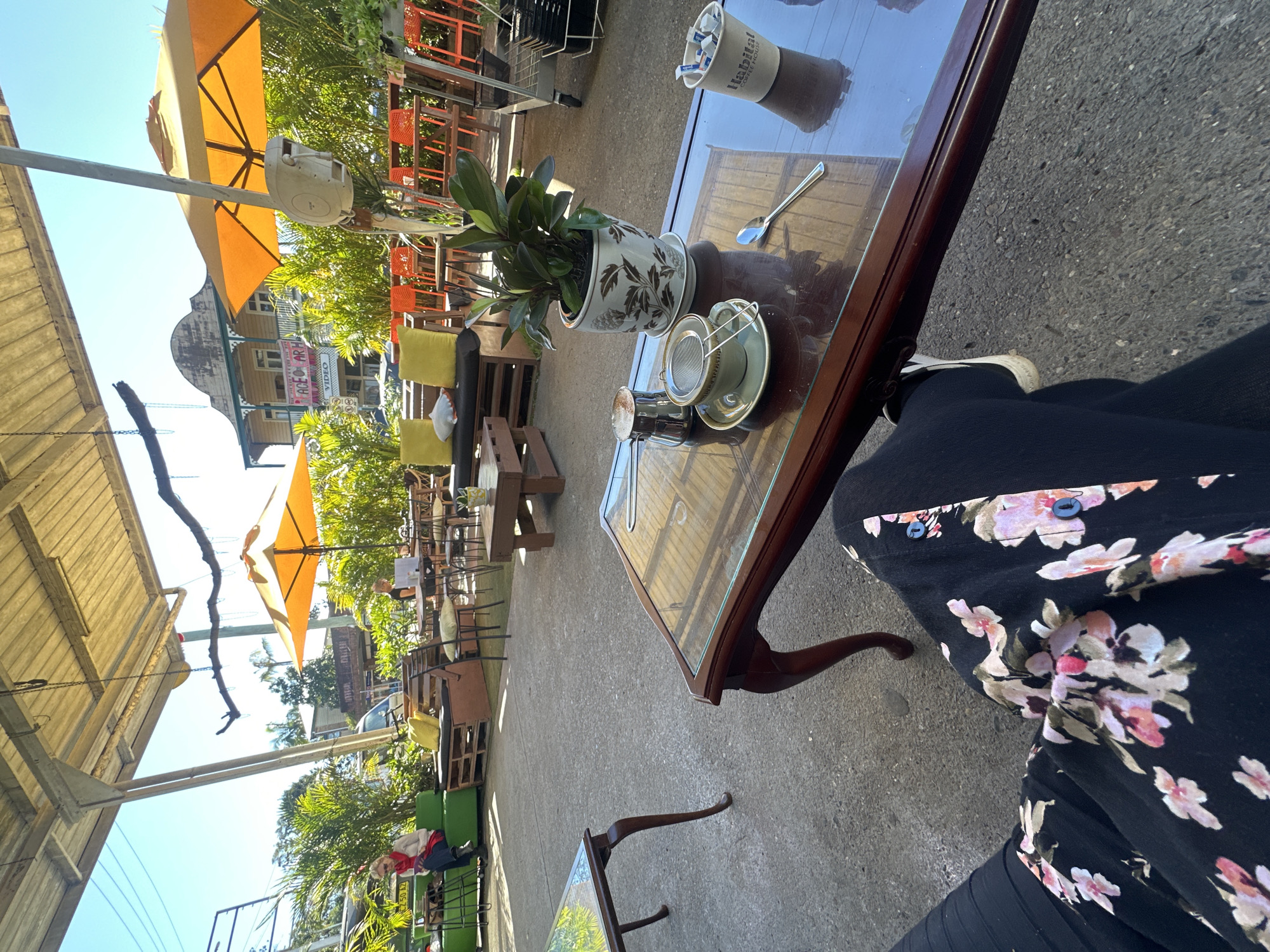 The image depicts an outdoor seating area of a cafe, likely in Bellingen, Australia, characterized by its relaxed ambiance and natural surroundings. In the foreground, a person's legs are visible, clad in black trousers with a floral pattern and white sneakers, seated at a dark wood coffee table with a glass top. On the table, there's a coffee cup with a strainer, a small metal pot, a spoon, and a bucket labeled "Habitat Coffee House" containing what appear to be sugar packets. A potted plant sits on the table, adding a touch of greenery.
The cafe's outdoor area features a covered patio with a metal roof and exposed beams, providing shade from the sun. Several tables and chairs are arranged around the space, with orange chairs visible in the background. Lush green plants and palm trees are scattered throughout the area, contributing to a tropical feel. In the middle ground, a woman is seated on a green outdoor sofa, reading a book. Further back, other patrons are seen at tables, some engaged with laptops, suggesting a common gathering spot. The background reveals a building with a distinctive architectural style, possibly a heritage building, with balconies and awnings. The presence of orange umbrellas and the overall outdoor setting indicate a warm, possibly sunny day, consistent with the climate of Bellingen. The scene conveys a sense of casual leisure and community, where people can relax, enjoy refreshments, and socialize.