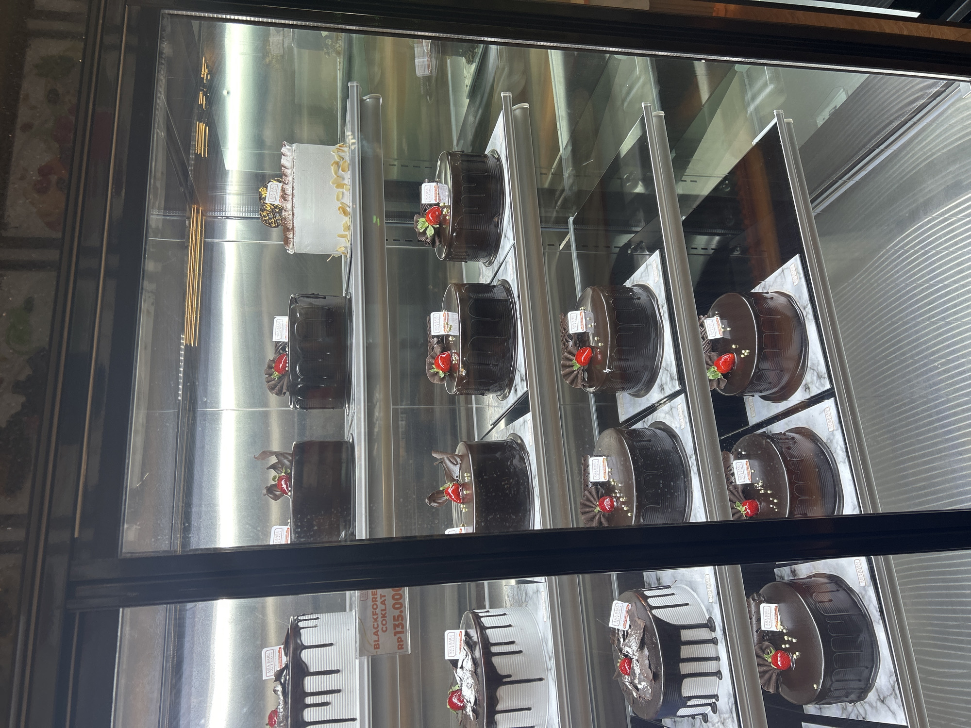 The image shows a display refrigerator filled with various cakes. On the top shelf, there are three cakes: a white cake with almond slivers and a small chocolate decoration on top, a dark chocolate cake with a decorative chocolate swirl and a strawberry on top, and another dark chocolate cake with a chocolate ribbon and a strawberry.
The second shelf from the top features four cakes. From left to right, there is a white cake with dark chocolate drizzles and a strawberry on top, a dark chocolate cake with chocolate drizzles and a strawberry on top, another dark chocolate cake with chocolate drizzles and a strawberry on top, and a partially visible cake on the far right. A price tag that reads "BLACKFORE COKLAT RP135.000" is visible below the first white cake on this shelf.
The third shelf from the top displays three cakes. On the left is a white cake with dark chocolate drizzles and a strawberry on top. To its right are two dark chocolate cakes, each with chocolate drizzles and a strawberry on top.
The bottom shelf shows two dark chocolate cakes, each with chocolate drizzles and a strawberry on top.
The cakes are displayed on marble-patterned plates within a refrigerated glass display case. The environment appears to be a bakery or cake shop. The lighting is bright, reflecting off the glass and the shiny surfaces of the cakes. The background inside the refrigerator is metallic and reflective. The image is taken from a slightly low angle, looking up into the display case. The overall impression is one of a well-stocked and appealing cake display. This scene is likely located in a bakery in Makassar, Indonesia, given the price indicated in Rupiah (RP).