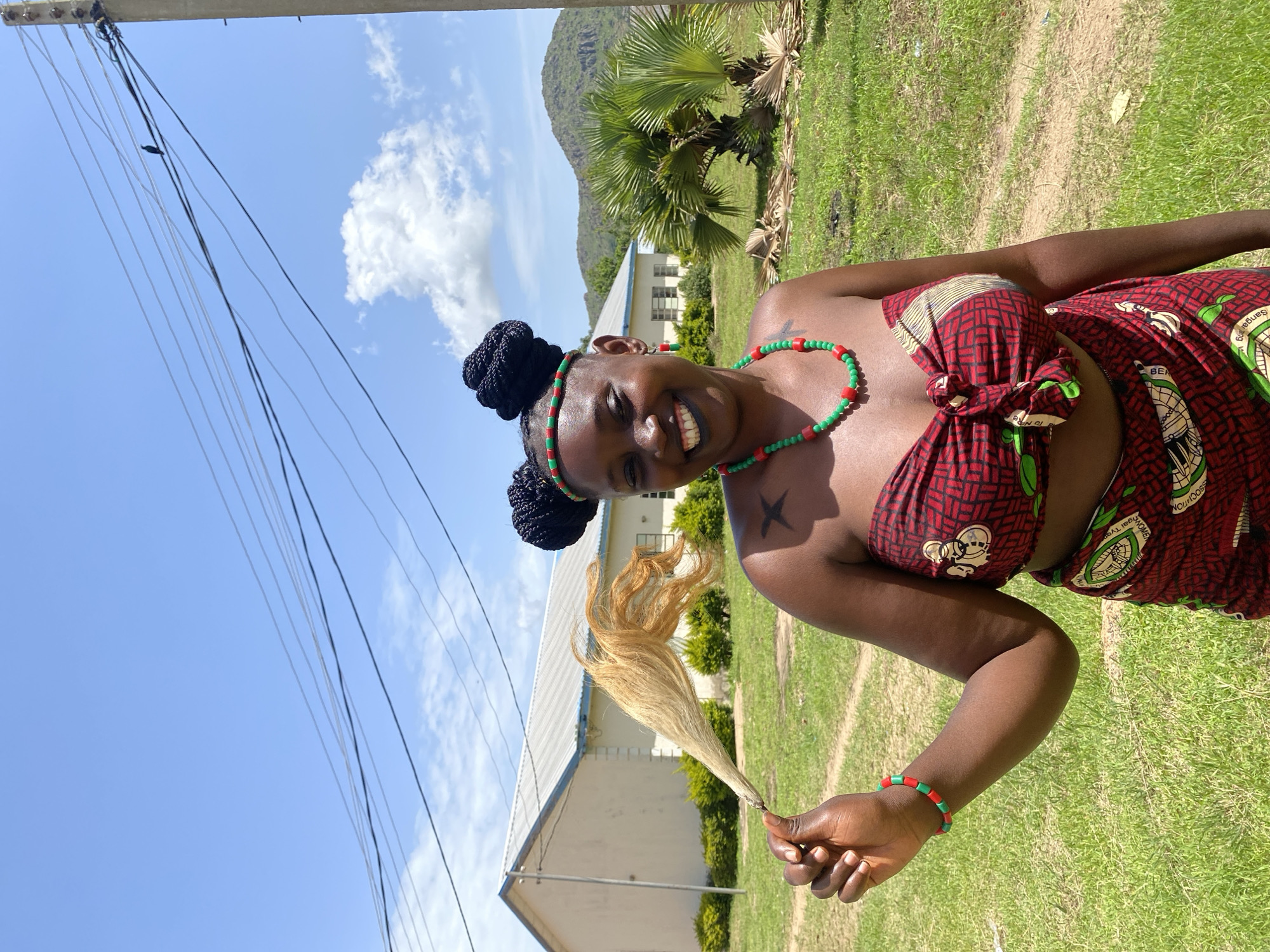 A young Black woman stands outside in what appears to be a sunny day with a clear blue sky and scattered clouds. She is wearing a red and black patterned wrap around her chest, and a matching patterned skirt. She is also adorned with red and green beaded jewelry, including a necklace and a bracelet. Her dark hair is styled in two buns on top of her head. She has a smile on her face and is holding a bundle of dried grass or straw in her right hand, which appears to be a whisk. There is a tattoo on her upper left arm.
In the background, there is a grassy area with some bushes and trees, including palm trees. A white building with a light-colored roof is visible behind her, and a hill with greenery can be seen further in the distance. A utility pole with several wires is in the left of the frame. The overall scene suggests a casual outdoor setting, possibly in a rural or semi-rural area, given the natural surroundings and the building. The weather appears pleasant, conducive to outdoor activities. The woman's expression and attire suggest she might be participating in a cultural event or activity, or simply enjoying a bright day outdoors. The location is identified as Vom, Nigeria.