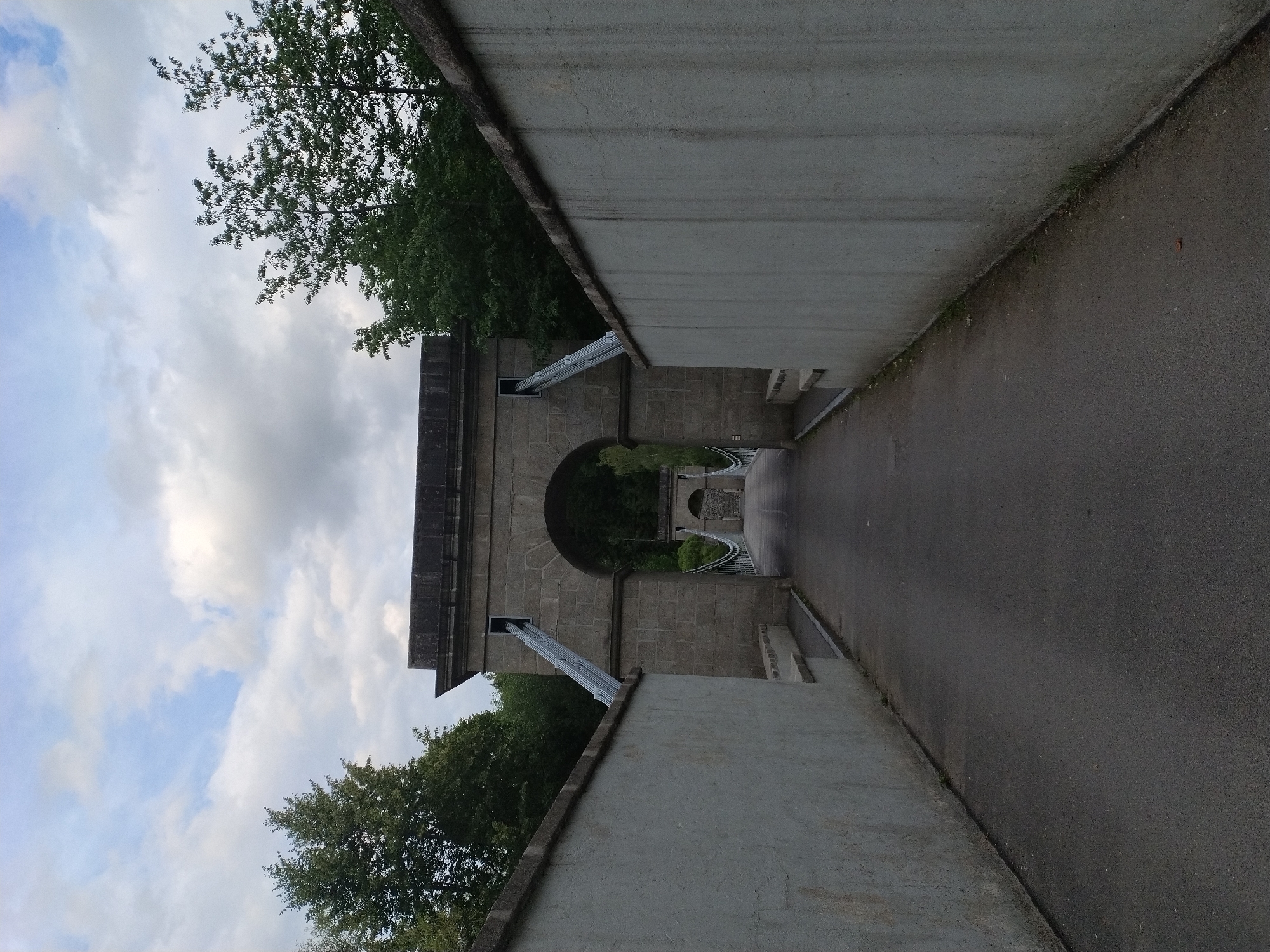 The image depicts a view looking down a paved pathway flanked by two substantial, light-colored concrete walls. The pathway leads towards an imposing stone archway, which appears to be part of a suspension bridge structure. Beyond the archway, a second, similar archway is visible in the distance, also part of the bridge. Cables from the suspension bridge are visible, extending upwards from the pathway and disappearing into the archway's structure. Trees line the background, with a cloudy sky above. The scene suggests a park or historical site. This is the Slavkovice suspension bridge, located in Slavkovice, Czechia.