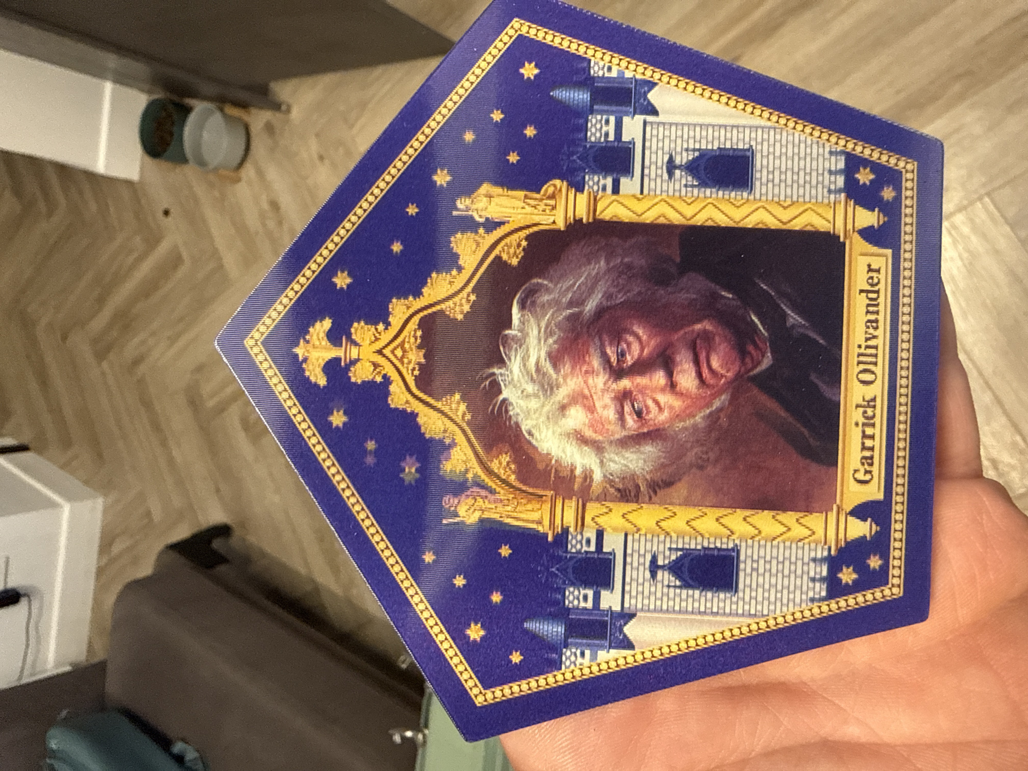 The image shows a Garrick Ollivander chocolate frog card from the Harry Potter series. The card is held in a person's hand. The background is a room with a light-colored herringbone pattern floor, a white cabinet, and a dark-colored piece of furniture. There are also two cylindrical objects, possibly containers, on the floor near the cabinet. There is no indication that this scene is located in Krasnoyarsk, Russia.