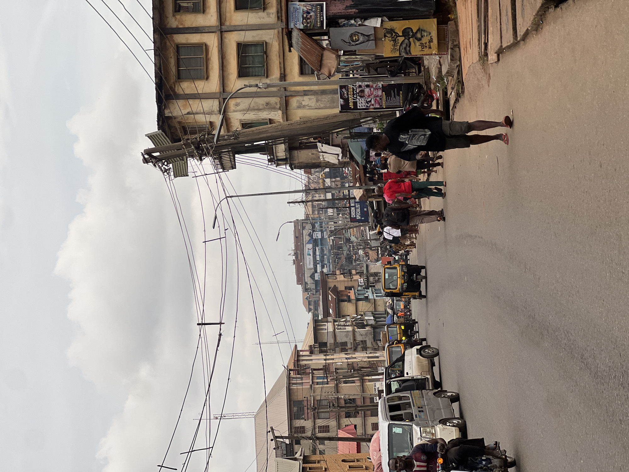 The image depicts a busy street scene in Nkpor, Nigeria. The perspective is from street level, looking down a paved road lined with multi-story buildings on both sides. The sky is overcast with visible clouds.
In the foreground, a man on a motorcycle is moving away from the viewer. To the left of the road, a white van is parked, and another white van is in motion. Further down the road, several yellow auto-rickshaws, a common mode of transport in Nigeria, are visible, some in motion and some parked.
Pedestrians are walking along the sidewalks and crossing the street. The buildings are a mix of styles and conditions, with some appearing weathered. Electrical wires crisscross above the street, supported by numerous utility poles, one of which is noticeably leaning. Various signs and advertisements are displayed on the buildings and poles. The overall impression is of a vibrant, active urban environment.
