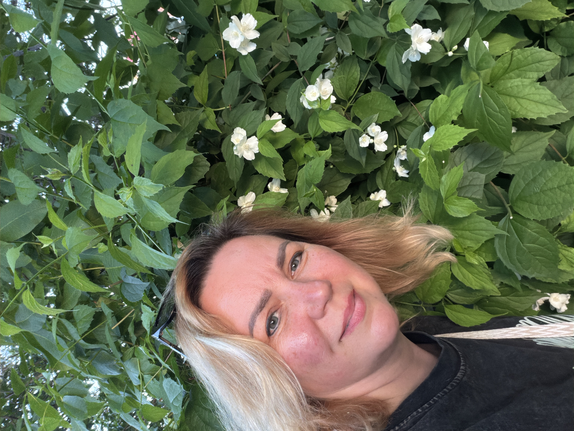 A woman with blonde hair is pictured in a close-up selfie against a backdrop of lush green foliage and white flowers. The location is suggested to be Leningradskaya, Russia, however, there is no specific evidence in the image to confirm this. She is wearing a dark-colored t-shirt. Sunglasses are perched to the side of her face. The dominant colors are green, white, and blonde.