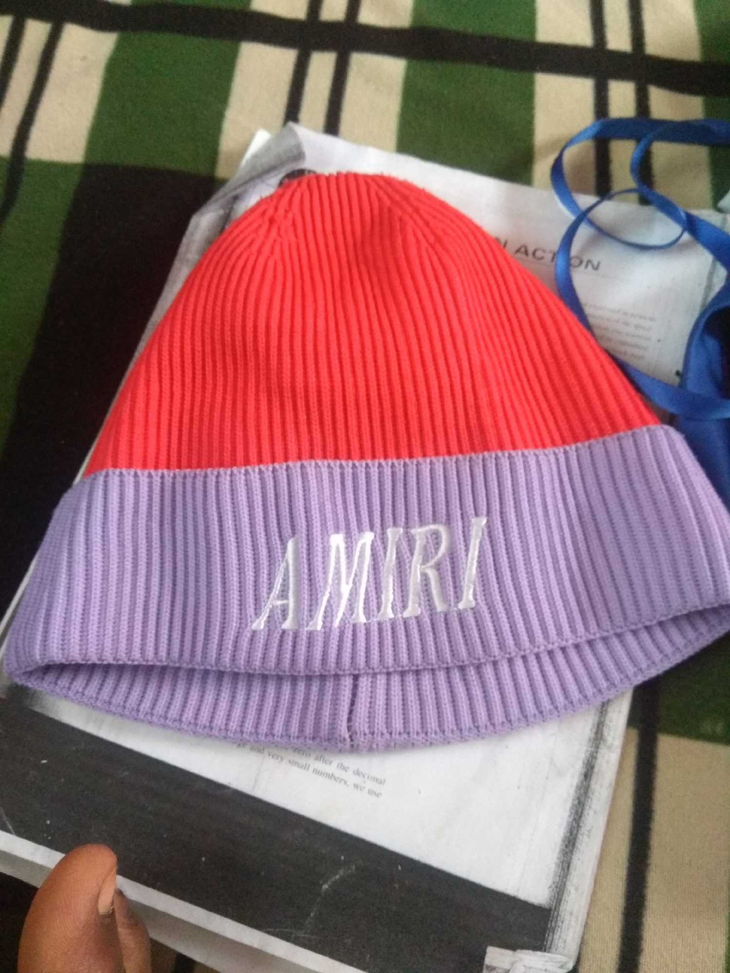 A beanie with "AMIRI" embroidered in white lettering on a lavender band is laid on top of a book. The beanie has a red upper portion and a ribbed lavender band. A blue ribbon is visible to the right of the beanie, partially obscuring text on the book. The book is resting on a striped green and beige cloth. The scene appears to be indoors. The image location is Jalingo, Nigeria.