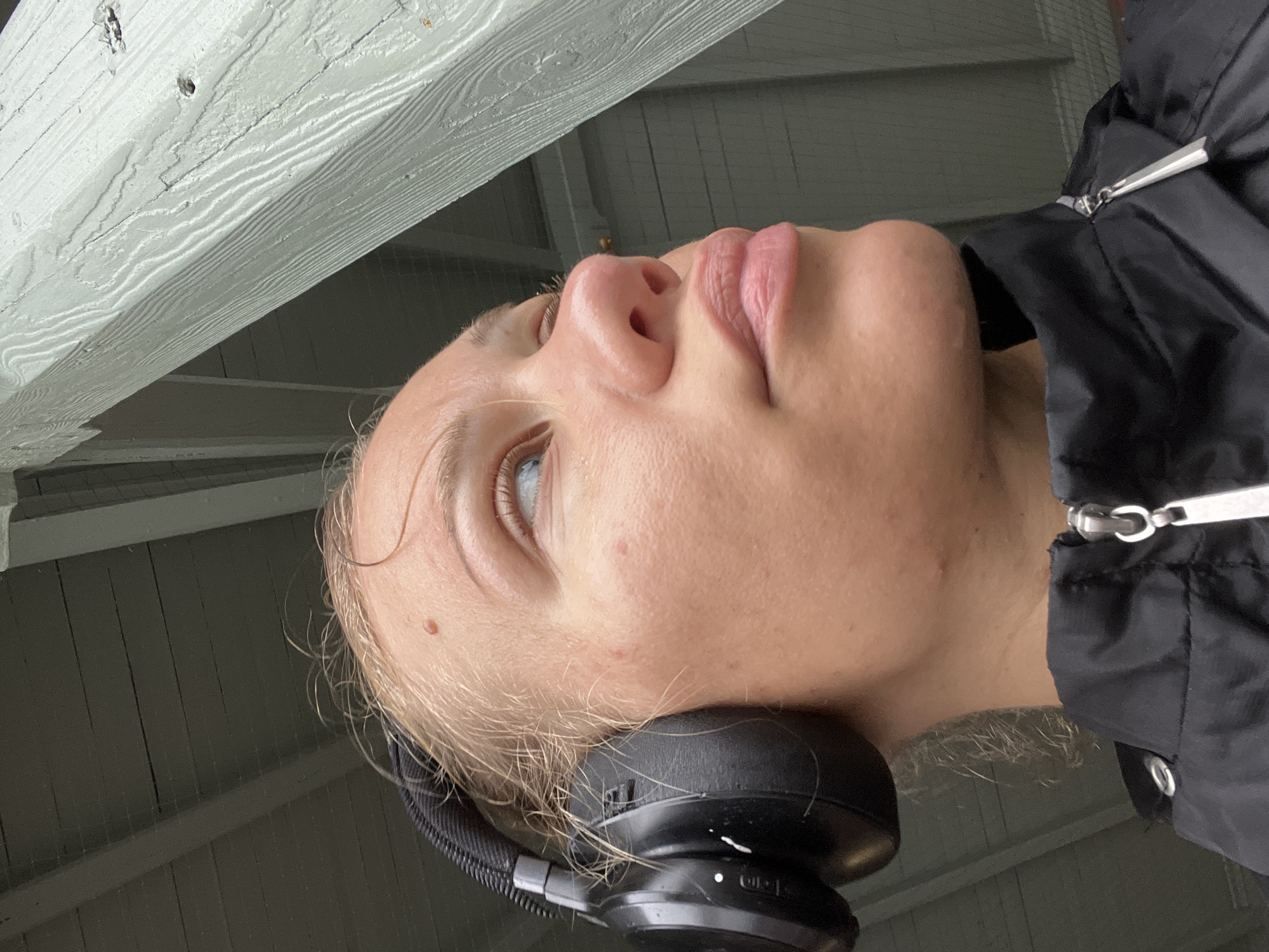 An outdoor, medium shot captures a close-up of a blonde woman wearing black headphones, looking upwards and to the right. Her face is illuminated by natural light, highlighting her skin texture, a mole on her forehead, and some minor blemishes. She is wearing a black zippered jacket. The background consists of a light green wooden structure, possibly part of a building or a covered outdoor area, with a dark ceiling of wooden planks visible above. The specific location in the city of Altötting, Germany, cannot be determined from the visual information provided.