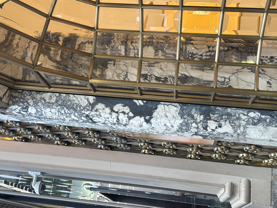 The image presents a close-up, upward-angled view of a building facade on Fifth Avenue in New York City, identified as the Cartier Mansion at 653 Fifth Avenue. The scene primarily details architectural elements.
To the left, a white, dome-shaped security camera is mounted on a light-colored stone wall. Below and to its right, a vertical section of a decorative gilded bronze grille features repeating, elaborate oval motifs. Adjacent to this grille is a prominent, vertically fluted column composed of highly veined white and dark gray marble. To the right, a large arched window with a brass-colored grid system divides the glass into multiple rectangular panes. The interior visible through this window is brightly illuminated with a warm, golden-orange light. Reflections of the marble column are visible in the window's glass. Further left, reflections in an unseen adjacent glass surface display blurred vertical lines and indistinct dark shapes, consistent with an urban street environment and other nearby structures. No individuals are directly visible in the immediate foreground, though reflections suggest an active urban setting.