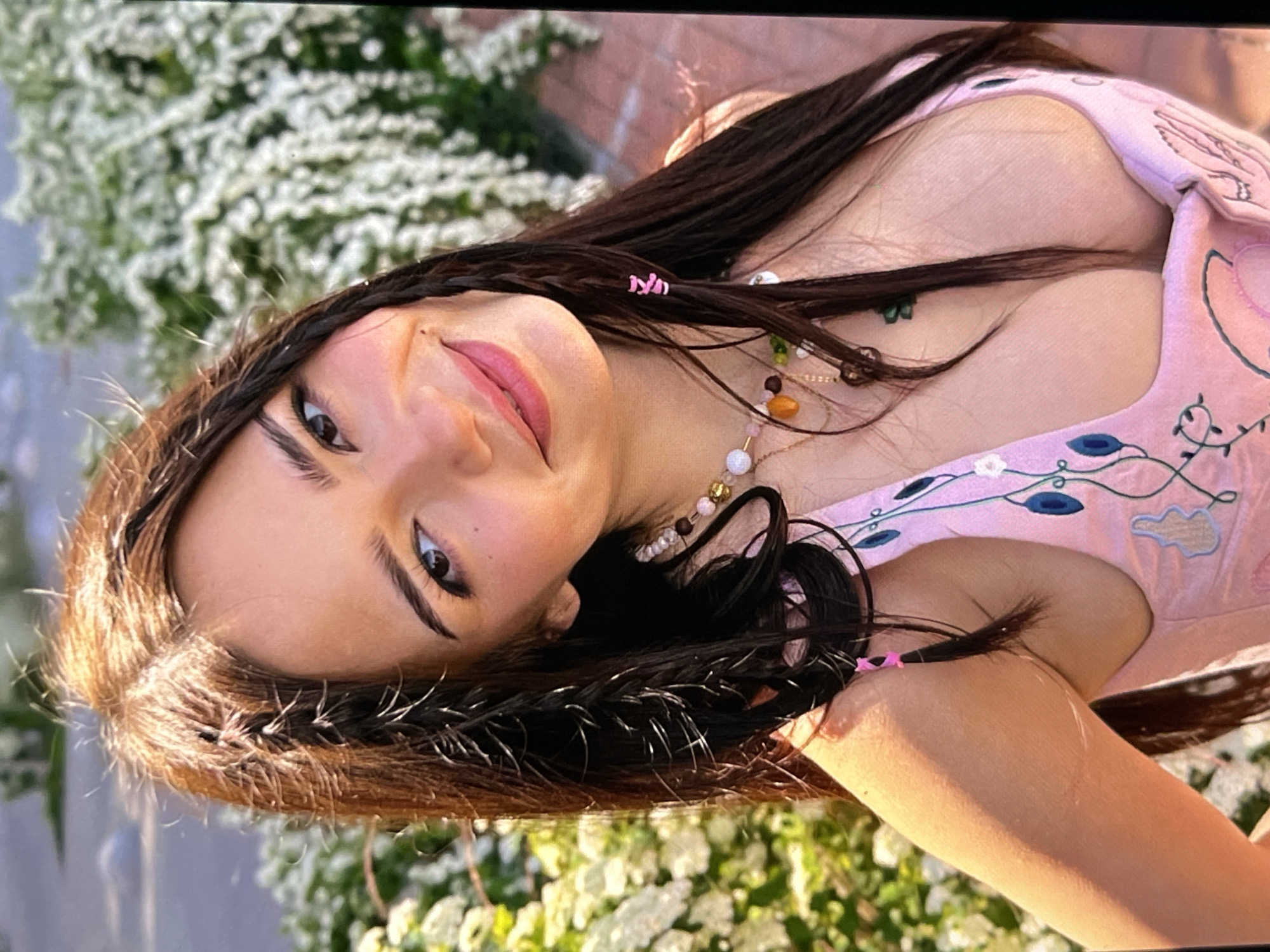 A young woman with long brown hair, styled with braids and pink hair clips, smiles at the camera. She is wearing a pink top with embroidered flowers and leaves. The background is blurred and appears to be an outdoor setting with greenery and white flowers.